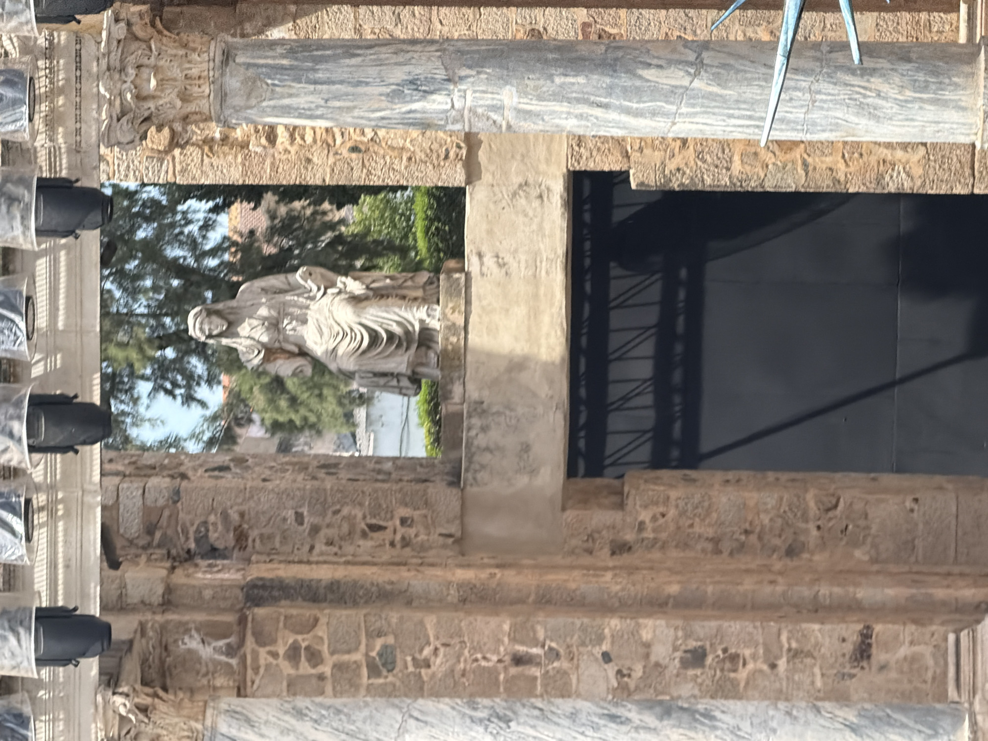 The image shows a section of the Roman Theatre of Merida, Spain. In the upper part of the image, several spotlights are visible. The central focus is a statue of a seated woman wearing a toga and a veil, positioned behind a stone archway. The statue is made of light-colored stone. To the left and right of the statue, there are stone columns. The left column is made of marble with grey veining, while the right column is also marble with grey veining. Behind the columns and the statue, there are trees and buildings. In the lower part of the image, a large dark door or entrance is visible, framed by stone architecture. Partially visible on the far right is a blue star-shaped sculpture.