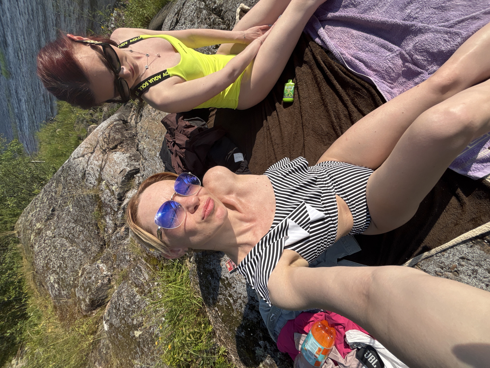 In this overhead shot, two women lounge on a rocky outcrop, bathed in bright sunlight. The woman in the foreground wears a black and white striped swimsuit and blue sunglasses. Her companion, positioned slightly behind her, is clad in a neon yellow swimsuit and dark sunglasses.
The setting appears to be a natural environment, likely near water, as suggested by the presence of rocks, grass, and a body of water visible in the upper left of the frame. A brown blanket and a purple towel are spread out on the rocks, serving as seating. Various objects are scattered around them, including a small green bottle, a black bag, and a bottle of orange liquid. The overall mood is relaxed and leisurely, indicative of a sunny day spent outdoors. The visible text on the neon yellow swimsuit straps reads "AQUA SOUL".