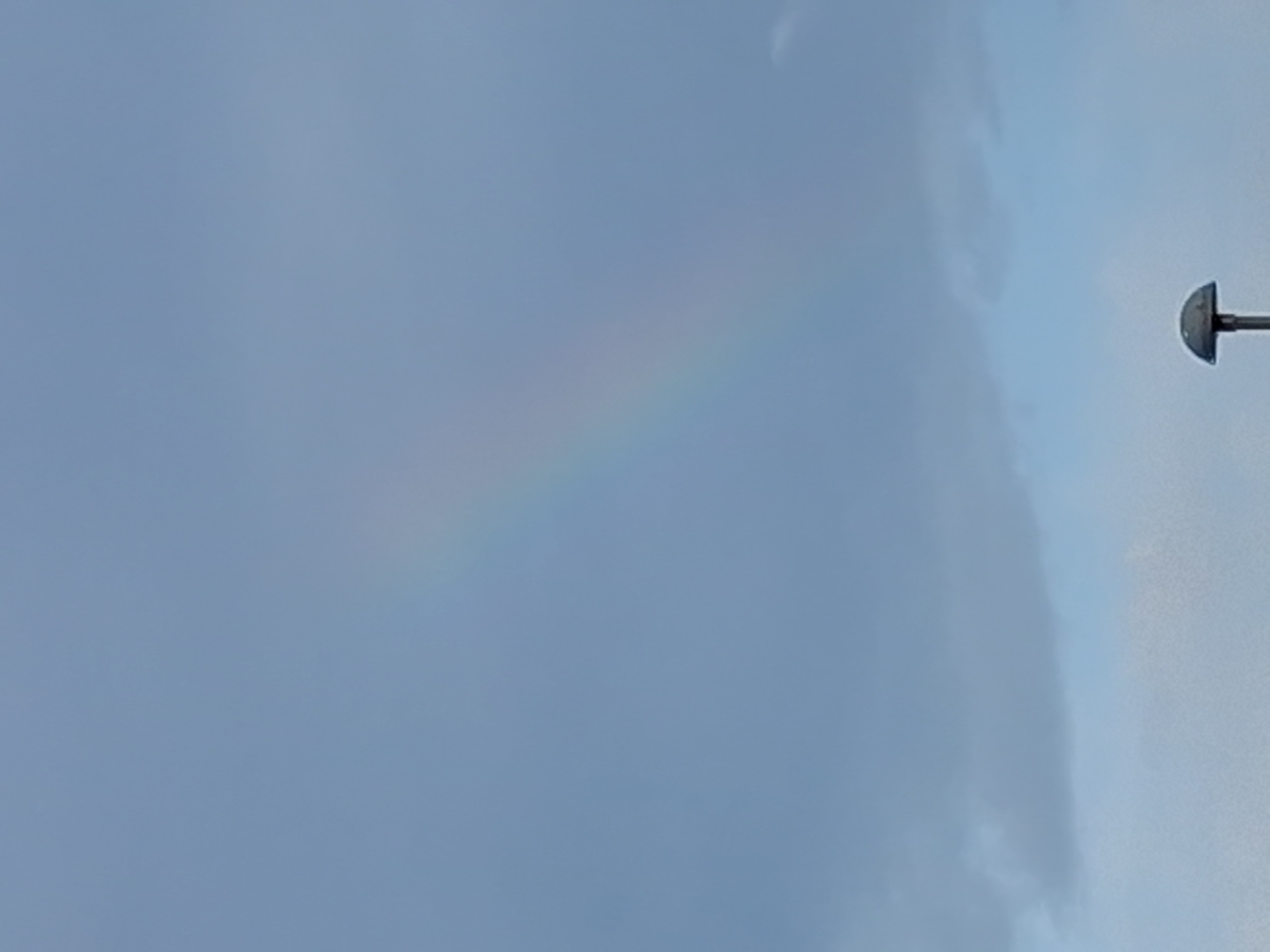 The image shows a pale rainbow in a cloudy sky. A street light fixture is partially visible at the edge of the image. The location in Figueira da Foz, Portugal is not discernible from the image.