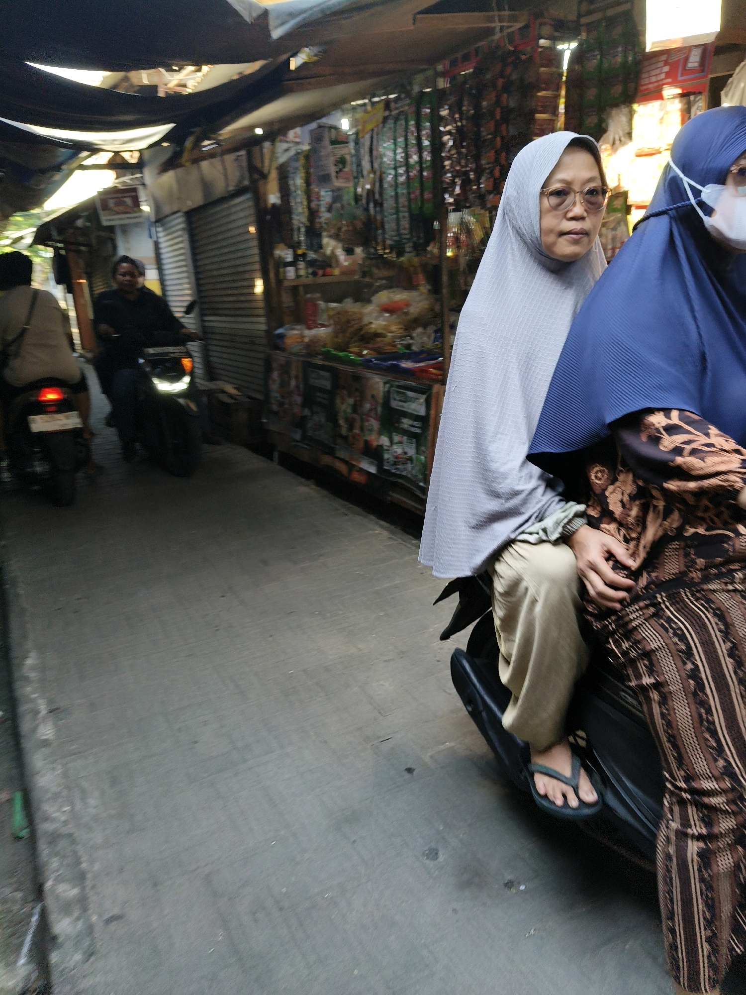 The image captures a street scene in Setiamekar, Indonesia, depicting a narrow lane lined with market stalls under makeshift awnings. In the foreground, two women are riding on a scooter. The woman in the front wears a light gray hijab and glasses, while the woman behind her wears a dark blue hijab and a face mask. The woman in the back is dressed in a patterned brown and black garment. They are captured mid-journey, with the scooter in motion on a paved surface. In the background, another person rides a scooter, and market stalls with various goods are visible, suggesting a bustling commercial area. The overall lighting suggests it is daytime.