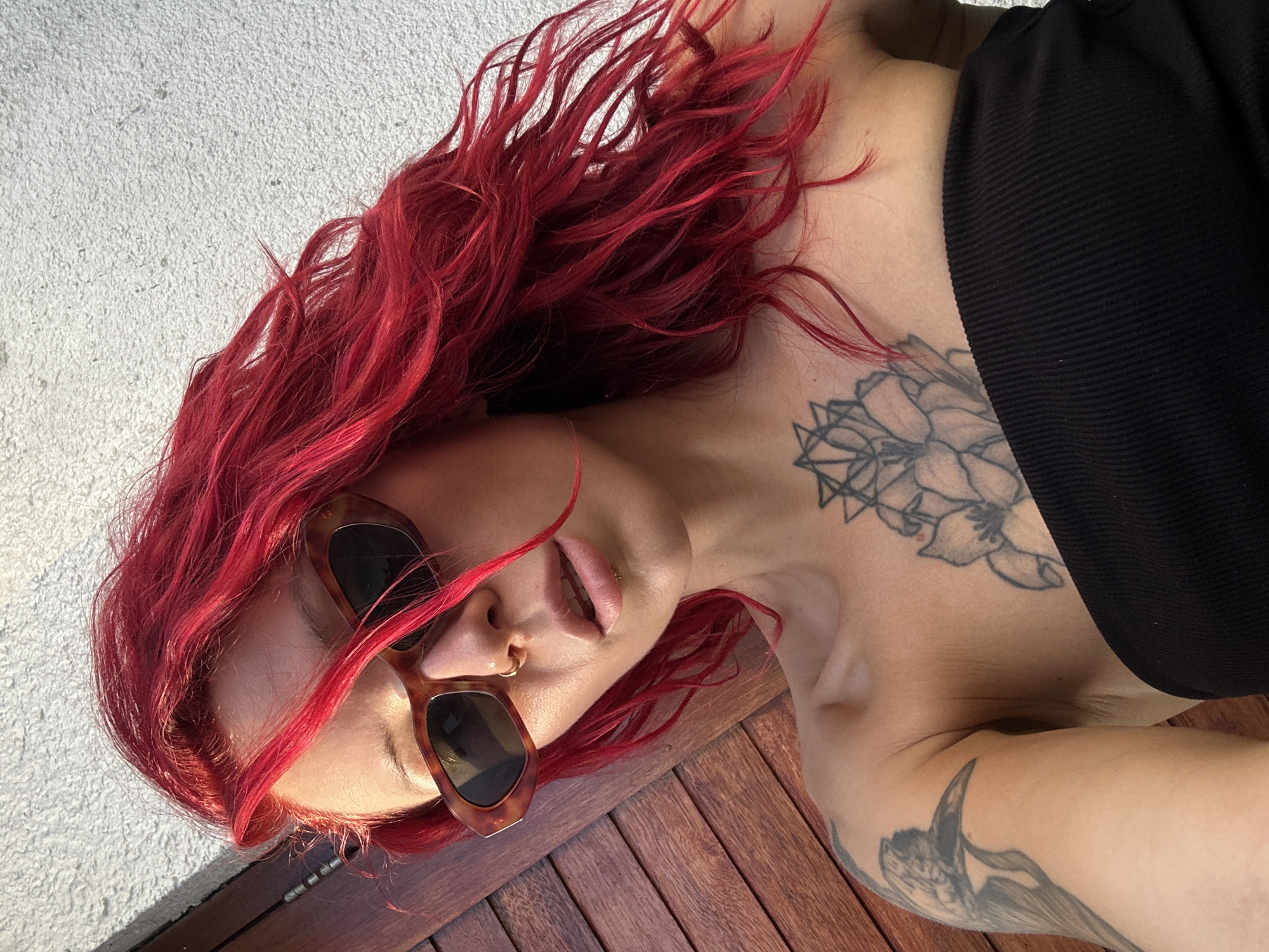 An individual with vibrant red hair lies down outdoors, seen from a high-angle perspective. The person is wearing dark brown, possibly tortoiseshell-framed, sunglasses and a black ribbed or pleated top. Their long, wavy red hair is spread out around their head. A small gold nose ring is visible on their right nostril, and a small stud adorns their upper lip, with their mouth slightly ajar.
Multiple tattoos are visible on the subject's upper body. A prominent floral or geometric design is inked on their left collarbone and chest area, and another tattoo, appearing to be a stylized bird or creature, is on their left arm.
The setting transitions from a light-colored, textured surface, possibly concrete or stucco, in the background to wooden planks beneath the person's head and arm. Strong, direct sunlight illuminates the scene, casting distinct shadows, indicating a bright, sunny daytime environment. The overall impression is one of relaxation or sunbathing. There is no visible text. The context places this scene in North Kuta, Indonesia.