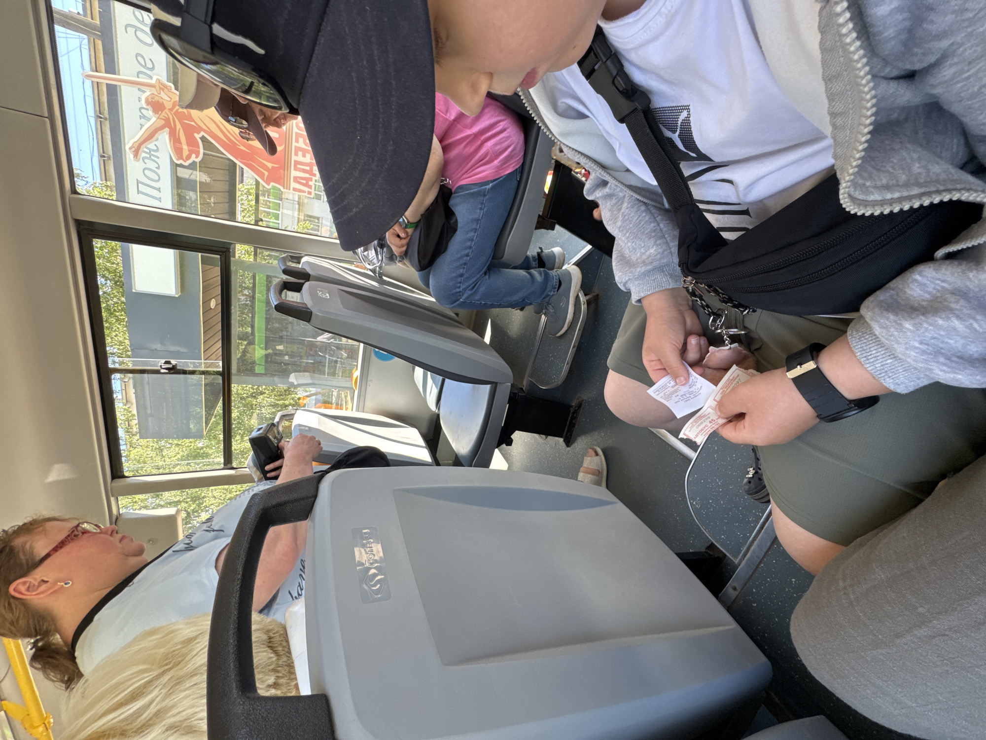 In Chaikovsky, Russia, inside a bus on a sunny day, a young person wearing a grey hooded jacket, a white t-shirt with a black graphic, and green shorts is holding two bus tickets. They are wearing a black wristwatch on their left wrist and a black fanny pack is slung across their chest. The bus interior features grey seats, a large grey plastic panel, and windows showing trees and a building with a large orange silhouette of a woman holding a sword and text that reads "Пожертвуй де". To the right, another passenger, an adult woman with glasses and her hair in a ponytail, is visible, holding a device and looking down. The overall scene depicts a mundane moment of bus travel.