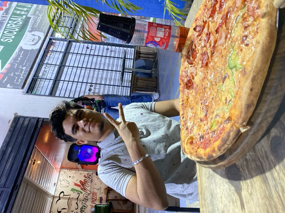 A young man holds up a peace sign next to a large pizza with pepperoni and green peppers, placed on a wooden board on a table. Behind him, another person stands by a window, and a sign reads "MAMMAMIA QUE PIZZA!" and "SUCURSAL #4.J". A black cup and a bottle of soda are also visible in the background.