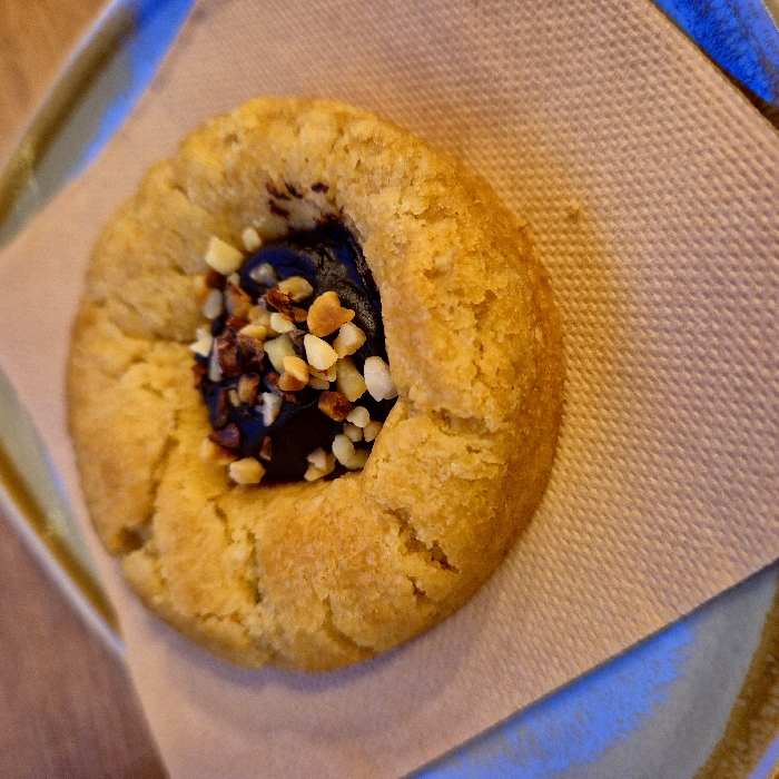 A close-up shot reveals a ring-shaped cookie resting on a beige napkin. The cookie has a golden-brown hue, and its texture appears slightly cracked. The center of the cookie is filled with a dark, possibly chocolate, substance, sprinkled with chopped nuts of varying shades. The napkin shows a subtle woven pattern. The overall image is warm-toned, with hints of blue from the surrounding plate.