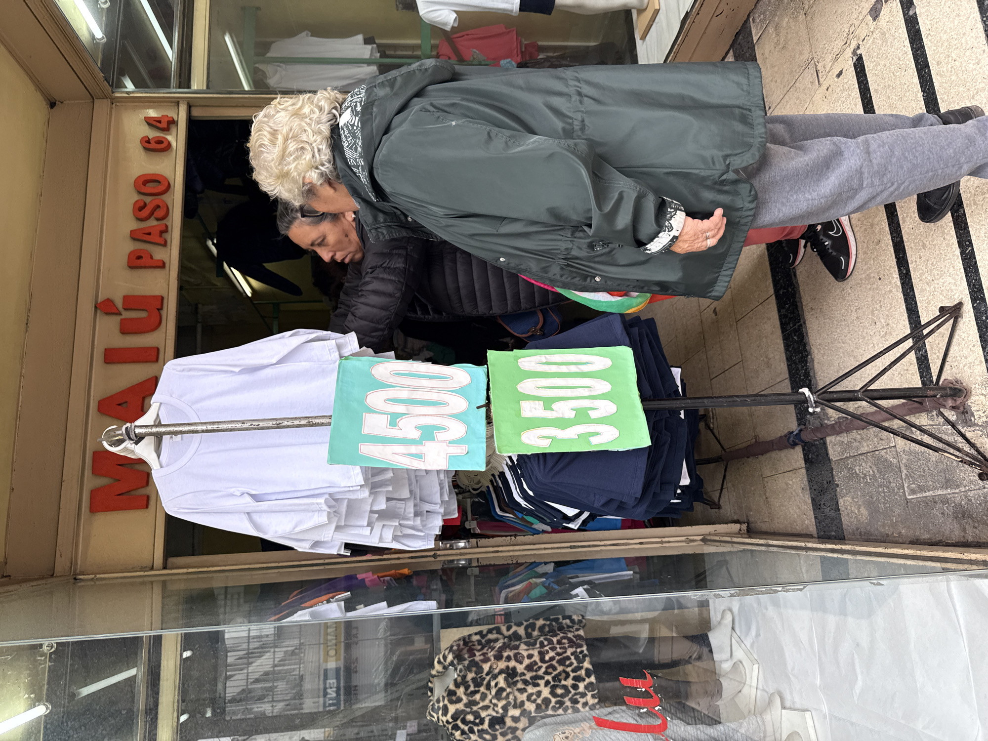 Here is a description of the image:
The image depicts a street scene in Buenos Aires, Argentina, specifically at a storefront located at "MaLú Paso 64." Two women are examining clothes displayed outside the shop. One woman has short, curly blonde hair and is wearing a green jacket. The other woman, with darker hair, is wearing a black quilted jacket. Clothes hang on a rack, with price tags reading "4500" and "3500" attached. In the storefront window, there is a reflection of various clothing items. The street surface consists of grey tiles and black stripes.