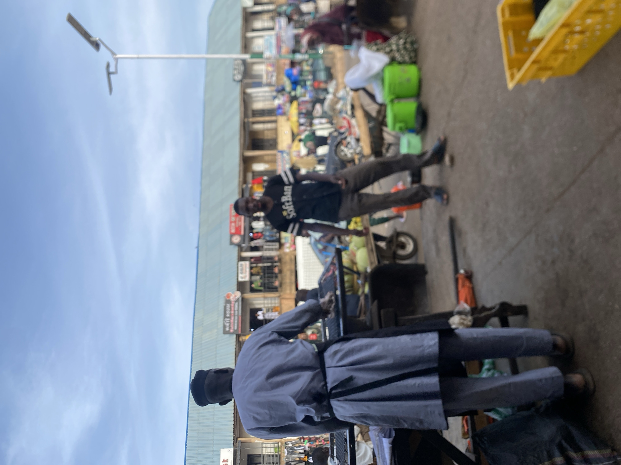 The image depicts an outdoor scene, possibly a street market in Bukuru, Nigeria. In the foreground, a man in a light grey robe and dark cap appears to be working at a grill. Behind him, another man is visible, wearing a dark shirt with "SoftBan" on it, along with khaki pants. A yellow basket is visible in the upper right corner of the image. The background shows a building with signs and multiple stalls, suggesting a busy marketplace. The sky is visible in the upper left corner. The scene is cluttered with various goods and people, a common characteristic of street markets.