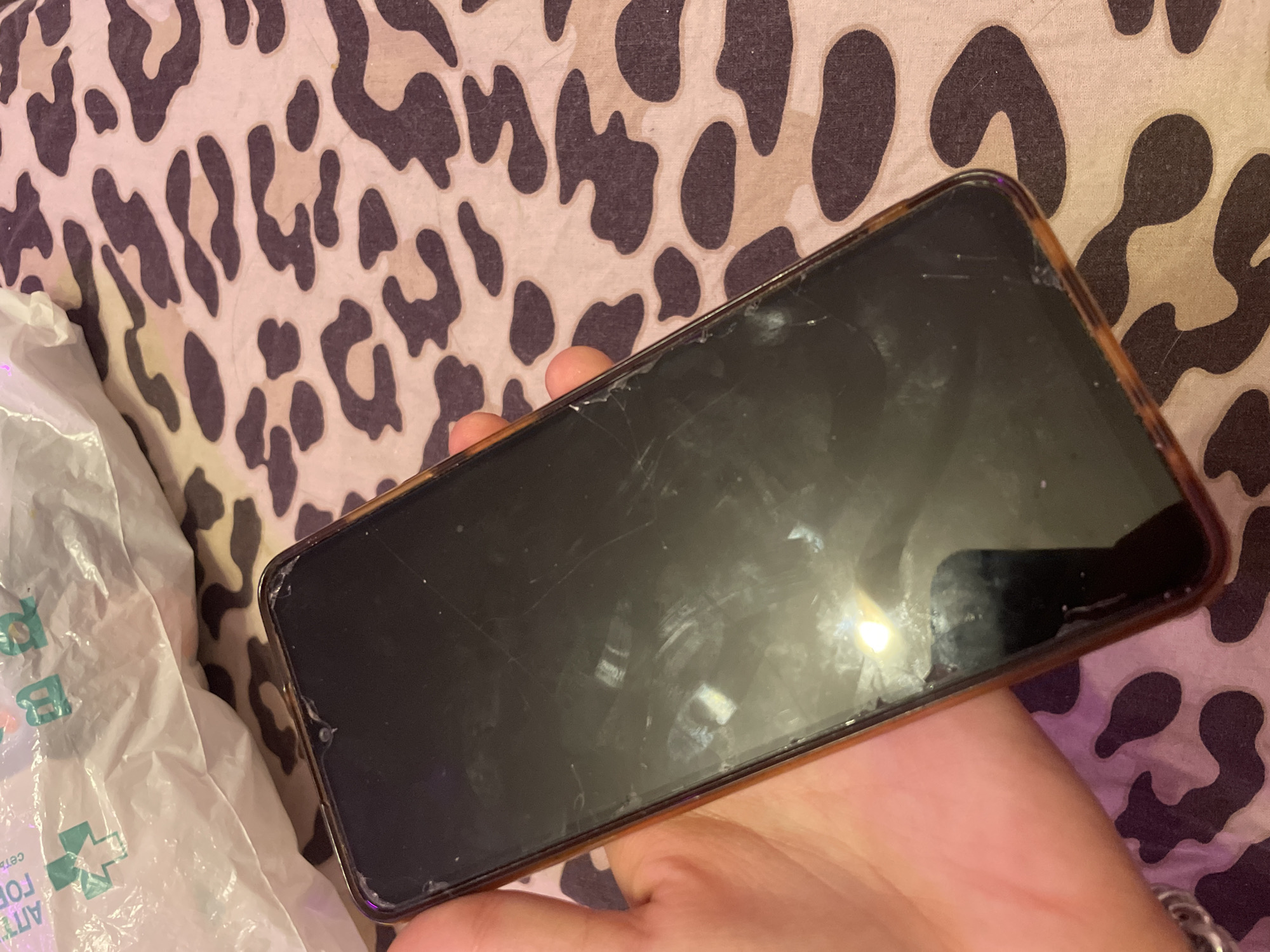 A hand holds a dark smartphone with a cracked screen, displaying multiple scratches and a smudge, against a leopard-print fabric background. A white plastic bag with green text and a cross symbol is partially visible in the upper left corner of the image. The lighting is dim, and reflections are present on the phone screen. There are no discernible landmarks or indications of the city of Irinovka, Russia.