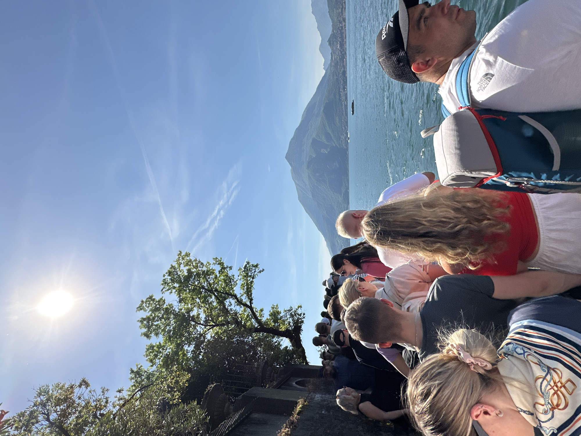 A large group of people are looking out over the water towards mountains under a blue sky with the sun shining. The scene is most likely set in Varenna, Italy, which is on Lake Como. The people are dressed in casual summer clothing and appear to be tourists. A man in a black hat and white t-shirt with a backpack is looking at the mountains on the right. A woman with long blonde hair, wearing a red top, stands behind him. A tree is present on the left.