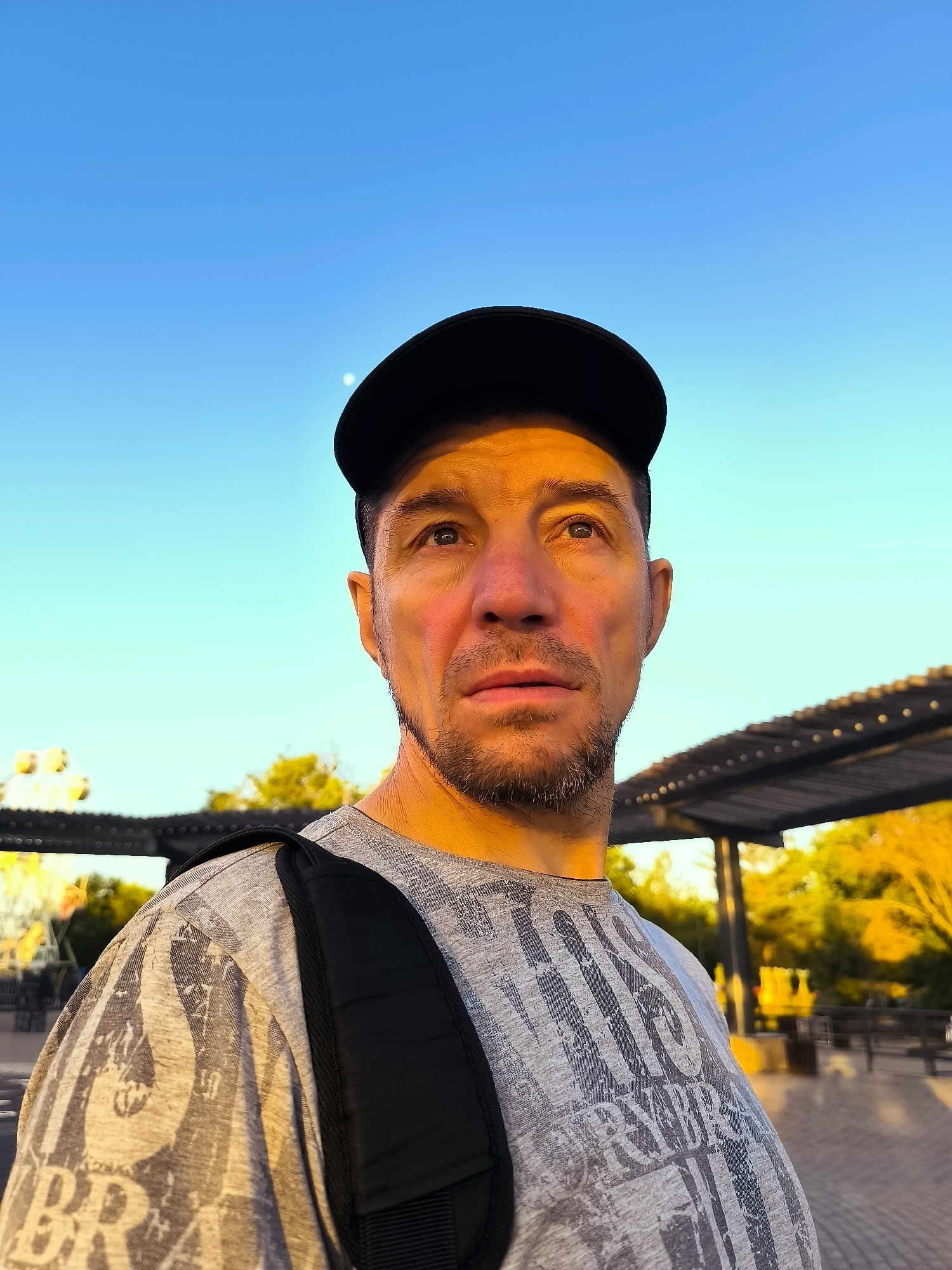 A man wearing a black baseball cap and a grey t-shirt with a graphic print is looking upwards and to the right. He has short, stubbly facial hair and appears to be in an outdoor setting. Behind him, a blurred background shows trees, a shaded structure with lights, and what appears to be a Ferris wheel in the distance. The sky is a clear, gradient blue, with a faint hint of the moon visible. The lighting suggests it is either early morning or late afternoon. There is no indication of the city of Qarağandı city administration, Kazakhstan.