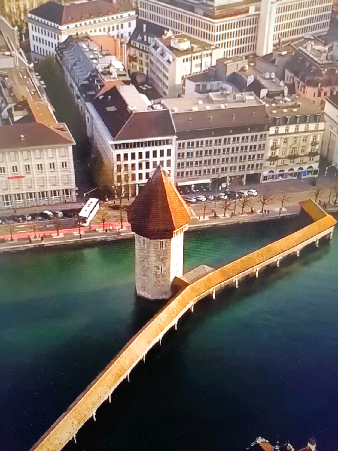 The image shows an aerial view of a medieval-style covered wooden bridge spanning a body of dark green water. The bridge has a reddish-brown tiled roof and connects to a stone tower with a similar roof. The tower appears to be hexagonal and is located in the water. Buildings with traditional European architecture, including a white building with many windows and a brown-tiled roof, line the waterfront. The scene is a city, likely in Europe, with several cars parked along the street by the water. There are no visible people or ongoing actions. The city of Netanya, Israel is not depicted in this image.