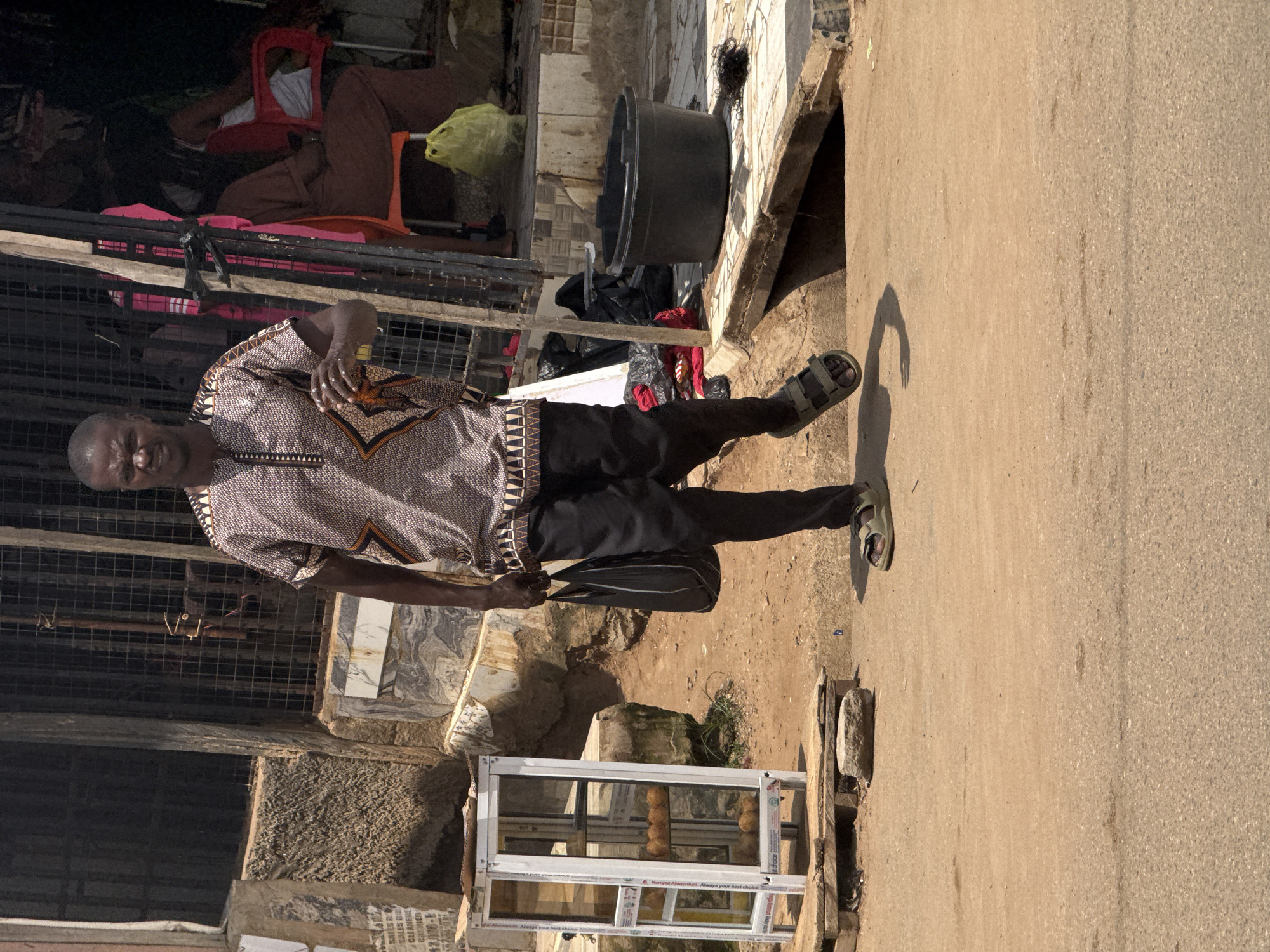 A man stands in an outdoor marketplace, facing the camera. He is wearing a traditional patterned dashiki shirt and dark pants, with sandals on his feet. He holds a black plastic bag in his right hand and appears to be looking to his left. His expression is neutral.
The setting is a bustling marketplace with stalls and structures made of metal, wood, and stone. In the background, there are racks and cages, suggesting a commercial area. A large black plastic tub is visible, along with bags of merchandise and what looks like food items in a glass display case. The ground is a mix of dirt and asphalt. Sunlight casts distinct shadows, indicating it is daytime, likely mid-morning or afternoon. The overall atmosphere appears to be casual and everyday.
Visible text includes what appears to be advertising slogans on the glass display case, such as "Hongtai Aluminium: Always your best choice." Other signs and labels are also visible but difficult to decipher. The location is identified as Abuja, Nigeria, suggesting an urban African environment. The weather appears to be sunny and warm.