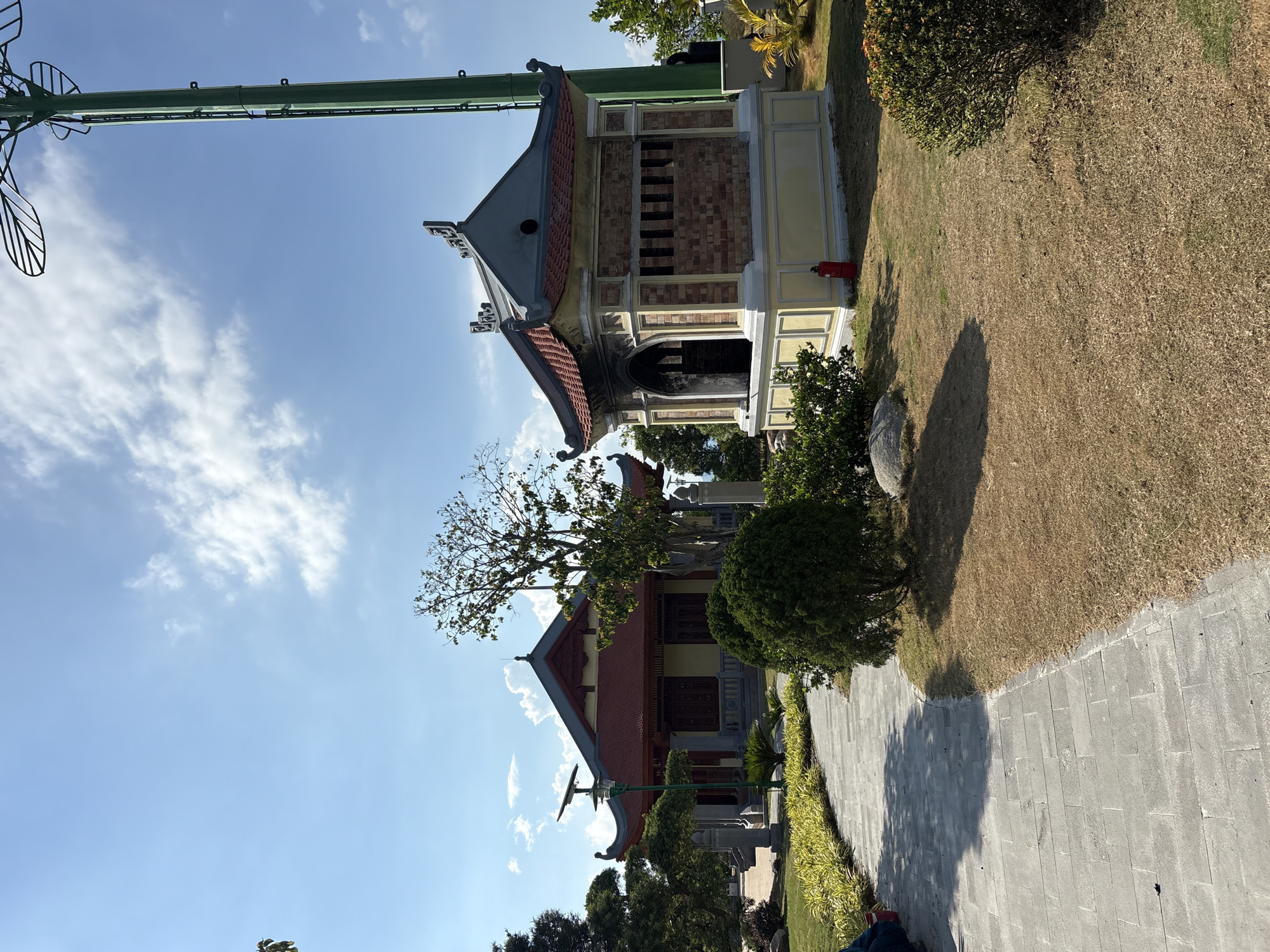 The scene is an outdoor setting in Tay Nha Trang Ward, Vietnam, featuring two traditional Vietnamese buildings under a partly cloudy sky.
The first building, closer to the foreground, has a red tiled roof, a yellow facade, and detailed woodwork around the doors. A tree partially obscures the front of the building.
The second building, behind and to the left of the first, is smaller with a grey roof and a tan facade. A fire extinguisher is visible near this building.
The buildings are surrounded by well-maintained landscaping, including neatly trimmed bushes and a dry, grassy area. A stone-paved pathway leads towards the buildings. A green metal structure (likely a lamp post or similar fixture) is visible on the left.