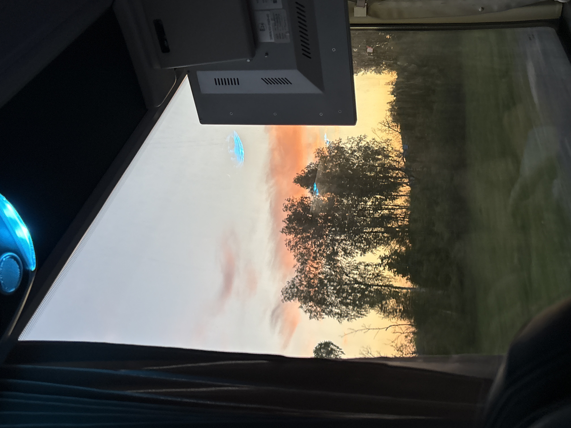 A view from inside a vehicle, possibly a bus, showing an overcast sky with a hint of orange and pink near the horizon, likely at sunset. Trees line the horizon, partially obscuring a field or open area. A monitor or device is mounted above, partially obscuring the view. The location might be Федькино, Russia, given the prompt context.