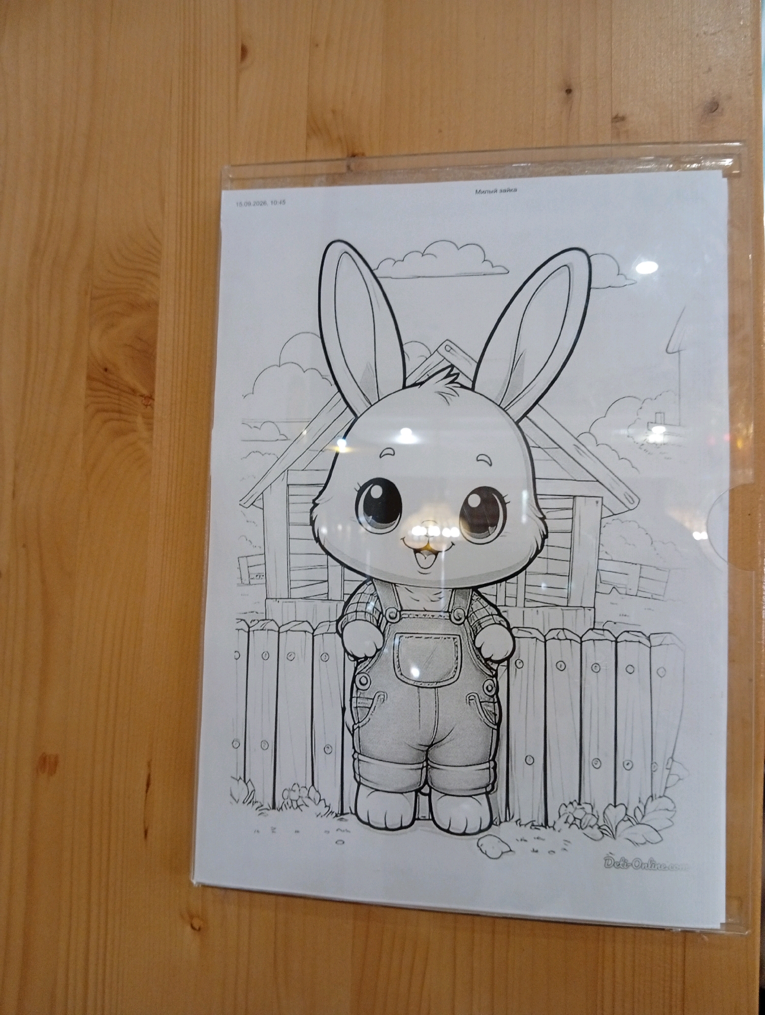 A black and white line drawing of a cartoon rabbit is displayed within a clear plastic sleeve, attached to a wooden surface. The rabbit, depicted in a naive, childlike style, wears overalls and a checkered shirt. It has large, round eyes, a small nose, and an open mouth with its tongue showing. Behind the rabbit, a simple illustration of a barn with a pitched roof and a wooden fence is visible. The scene appears to be a coloring page, with the text "Милый зайка" (meaning "Cute bunny" in Russian) and a date "18.09.2026, 10:45" printed at the top. A watermark or website address "Detsi-Online.com" is present at the bottom right of the drawing. The wooden surface suggests the image is displayed indoors, possibly on a wall or door, in a location that has not been identified.