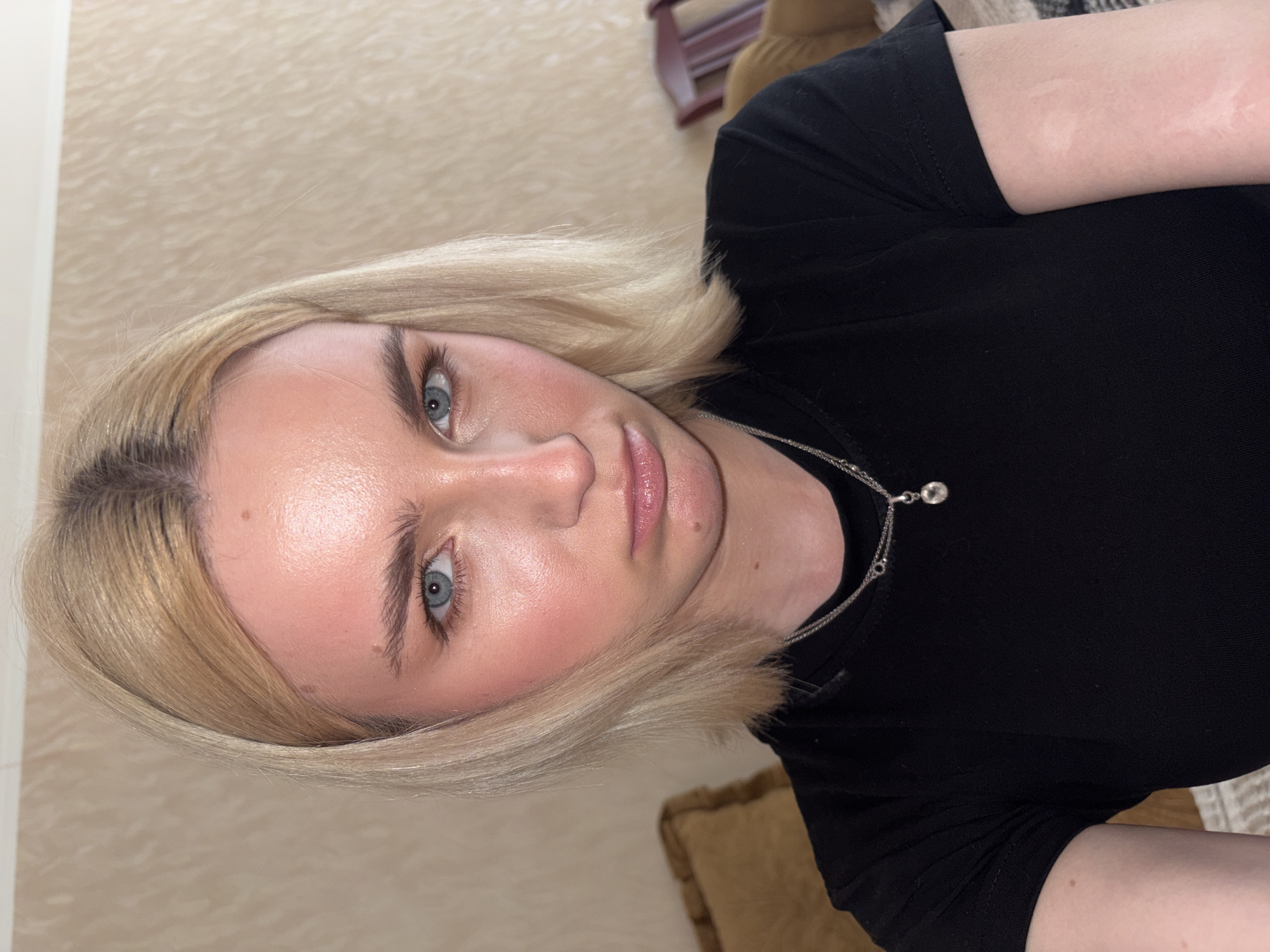 A young blonde woman with light blue eyes and fair skin, wearing a black t-shirt and a silver necklace with a pendant, is looking directly at the camera. Her expression is neutral, with slightly pursed lips. The background is an indoor setting with a textured wallpaper and furniture. The image was taken in Sinyavino, Russia.
