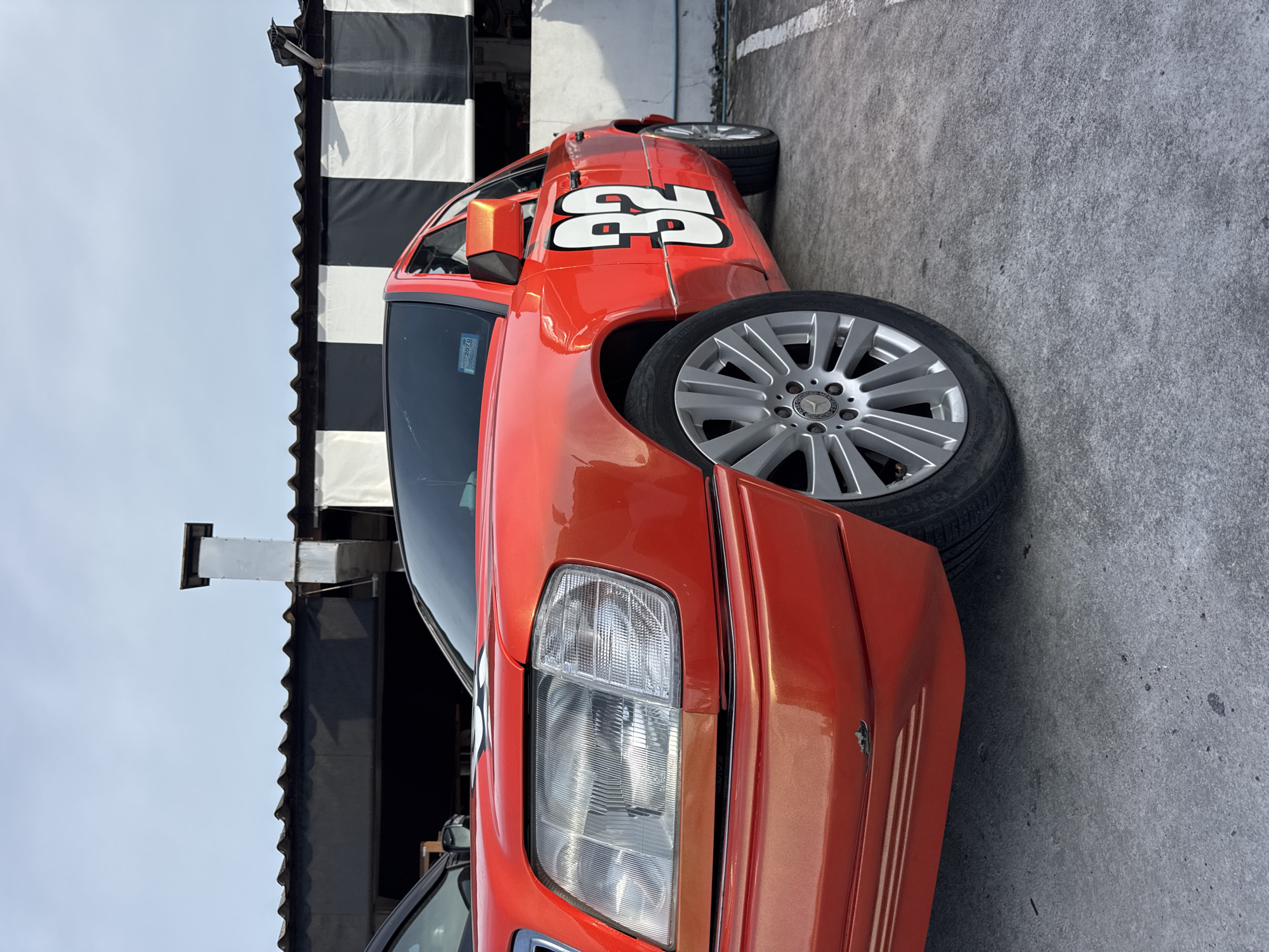 A bright orange Mercedes-Benz sedan with the number "32" in white on a black background is parked on a concrete surface in front of a building with a black and white striped facade. The car is angled towards the camera, showing its front passenger side. The building appears to be a commercial establishment, possibly a restaurant or shop, with a corrugated roof and a metal exhaust pipe protruding from it. The sky is overcast. There are no people visible in the image. The scene is located in Koh Phangan, Thailand.
