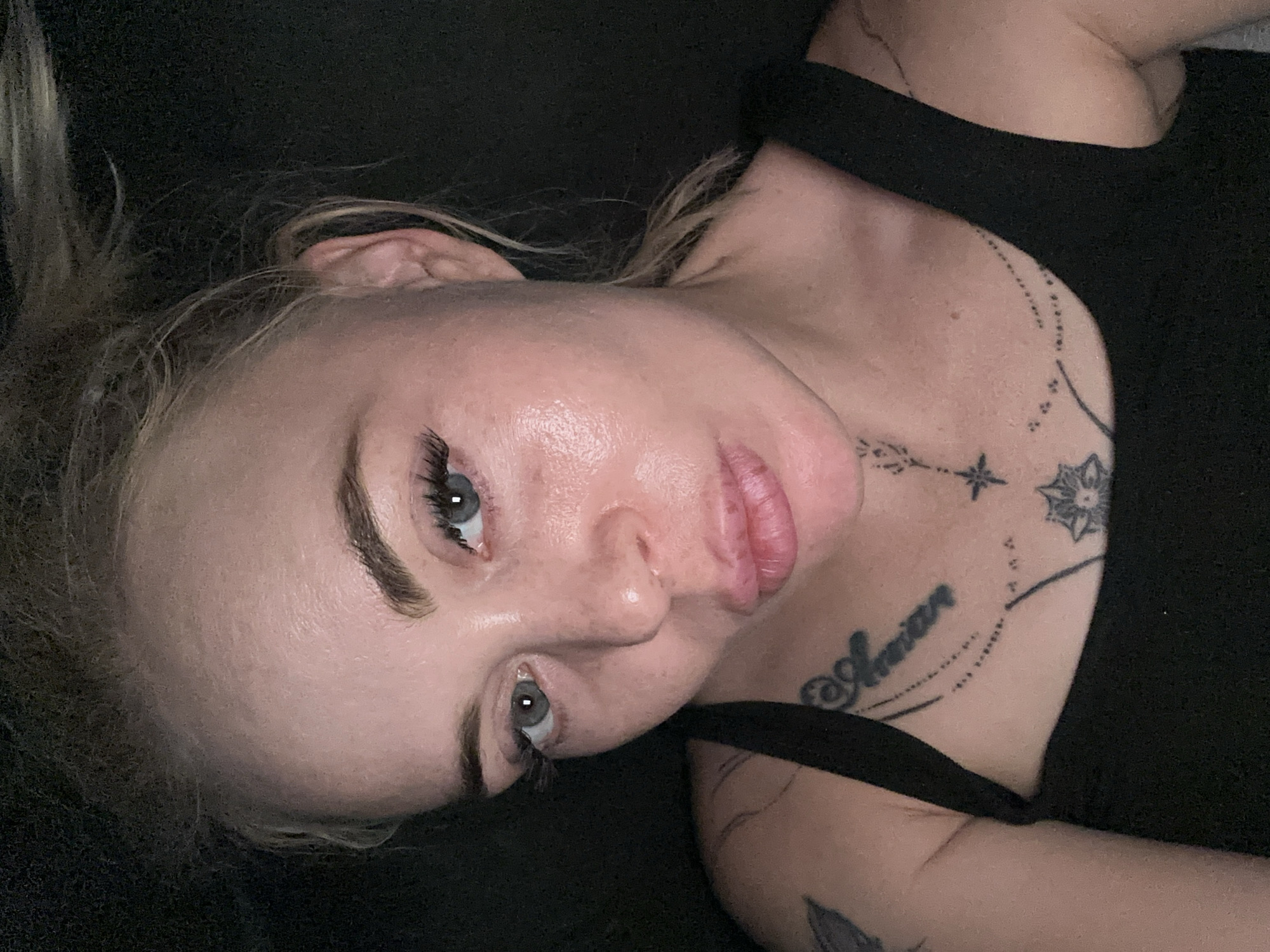 The image is a close-up, high-angle, selfie-style photograph of a woman's face and upper chest. The woman has blonde hair pulled back loosely, exposing her forehead, ears, and the sides of her face. Her eyes are open and looking towards the camera, with visible eyelashes and eyebrows. Her skin appears to be slightly oily. She is wearing a black tank top, and there are tattoos visible on her chest and left shoulder. The background is dark and out of focus. The image appears to have been taken indoors with artificial lighting. There are no visible indications of the location being in a city.