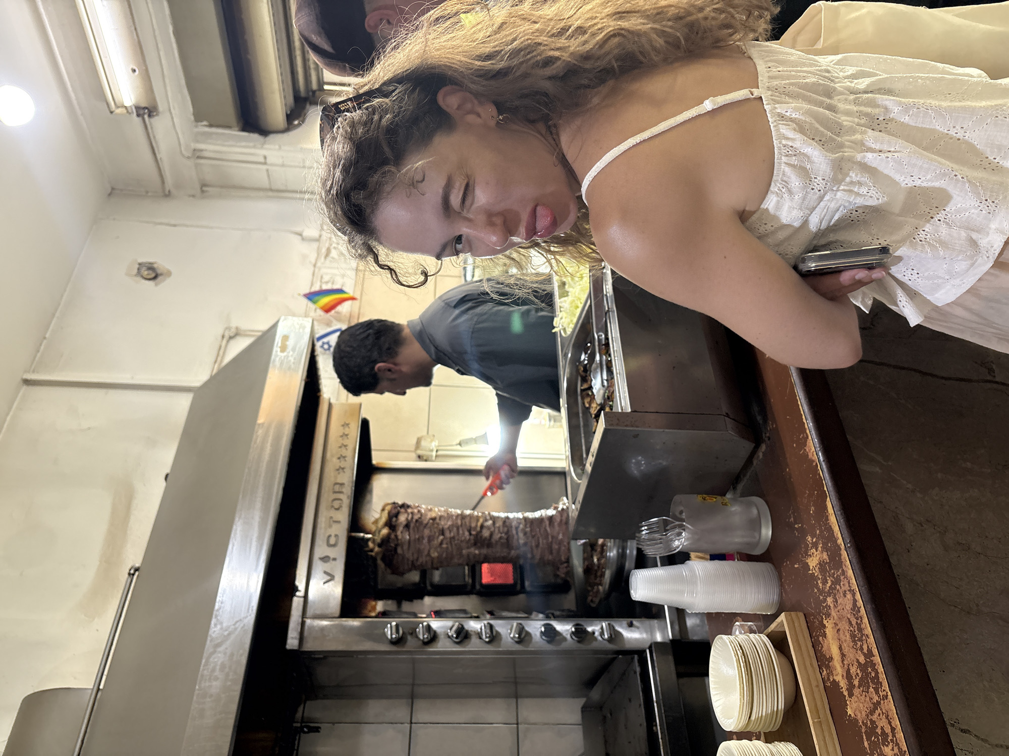 In an indoor food establishment in Tel Aviv, Israel, a woman with curly brown hair, wearing a white embroidered camisole, is captured in a close-up shot from the chest up. She is winking and sticking her tongue out, looking over her shoulder towards the camera. Her left arm is crossed over her chest, holding a smartphone. In the background, a man in a dark shirt is working at a shawarma spit, using a red tool to trim the meat. A vertically rotating spit of meat is prominently featured in the center of the frame, lit by red heating elements. Behind the shawarma machine, a small Israeli flag and a rainbow flag are displayed on the wall. The scene appears to be a busy eatery with food preparation stations and service counters visible. The counter in the foreground is made of dark wood and displays stacked small bowls, plastic cups, and cutlery.