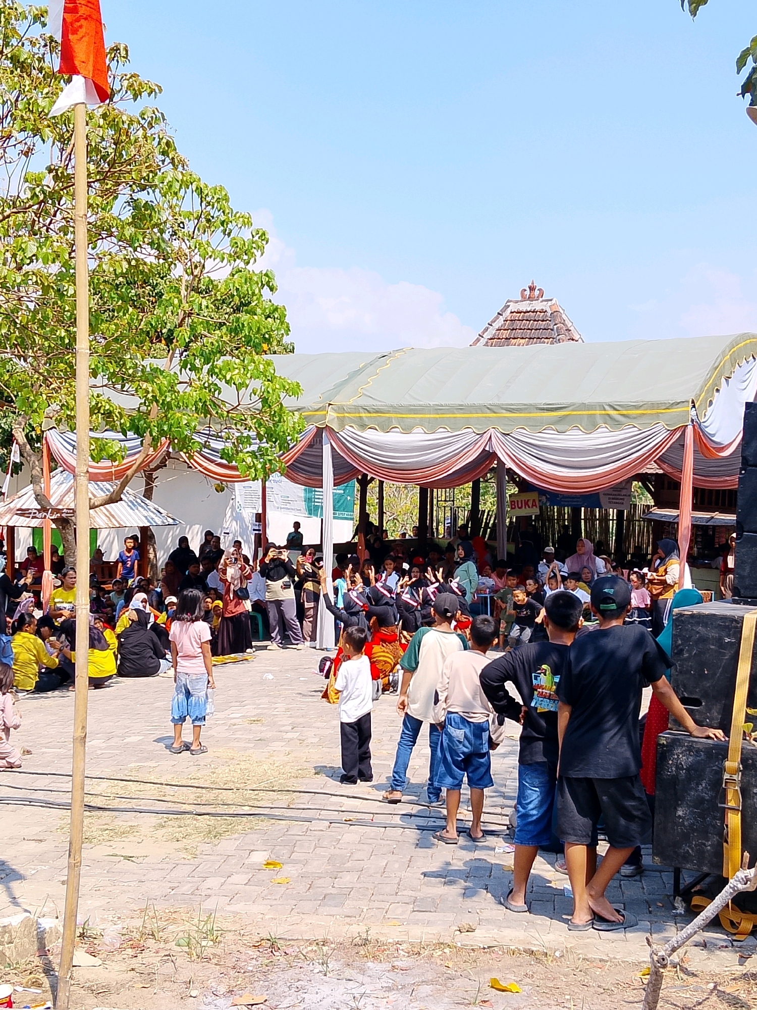 The image depicts an outdoor gathering in Ngaglik, Indonesia, likely a community event or celebration. A large tent with red, white, and grey draping covers a stage or performance area. In the foreground, several children and adults are present, some standing and others seated on the ground. A bamboo pole with an Indonesian flag is prominently displayed on the left side of the image. A large sound system, including speakers, is visible on the right. The ground is paved with interlocking bricks. In the background, more people are gathered, and the roofs of some buildings are visible, including a traditional Javanese-style roof. The sky is clear and blue. The overall atmosphere appears to be festive and social.