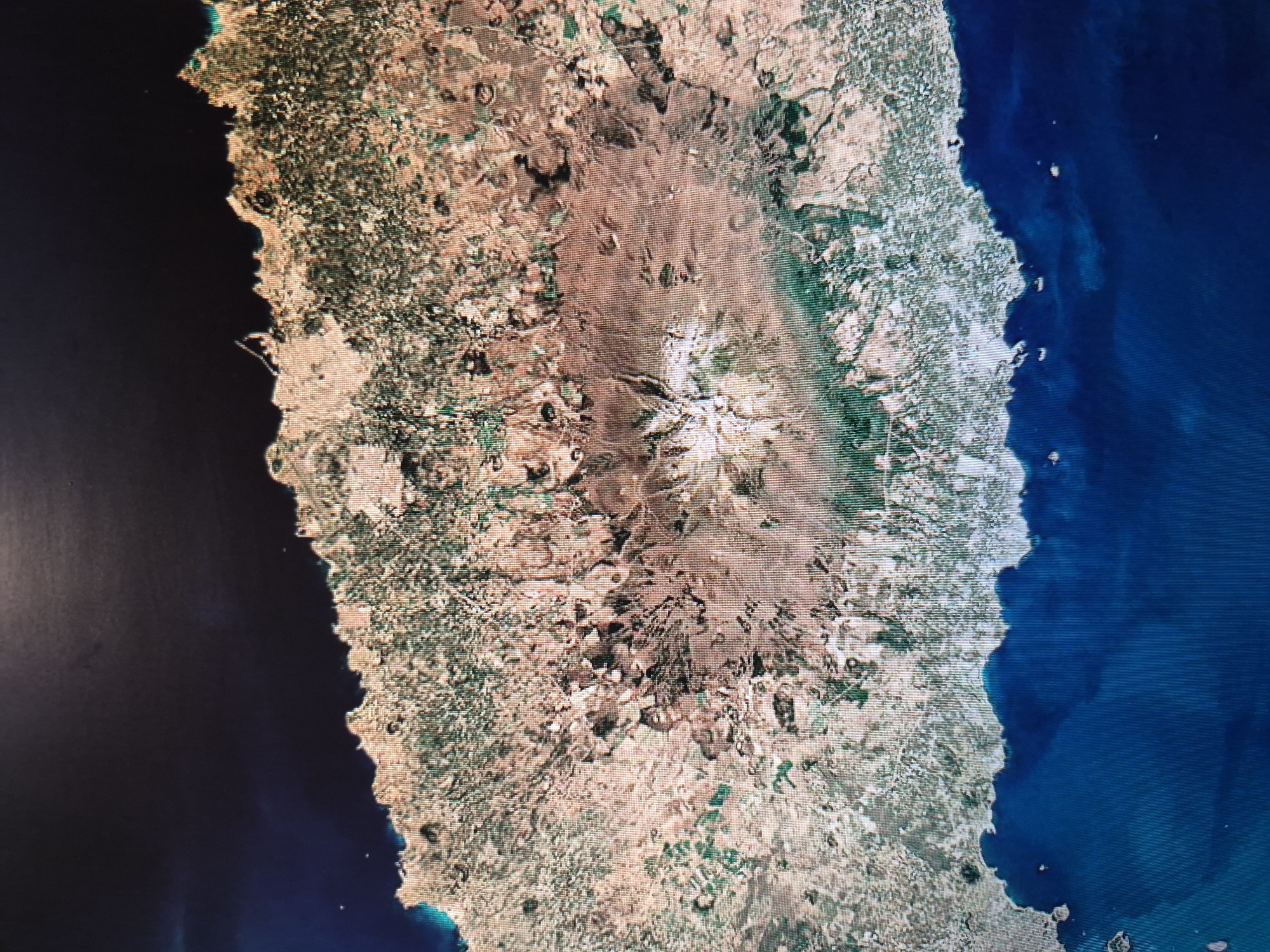 This is an aerial view of Mount Etna in Sicily, Italy. The image shows the volcanic mountain and surrounding landscape, including areas of vegetation, barren land, and the adjacent coastline with the Ionian Sea on the right and the Tyrrhenian Sea on the left.