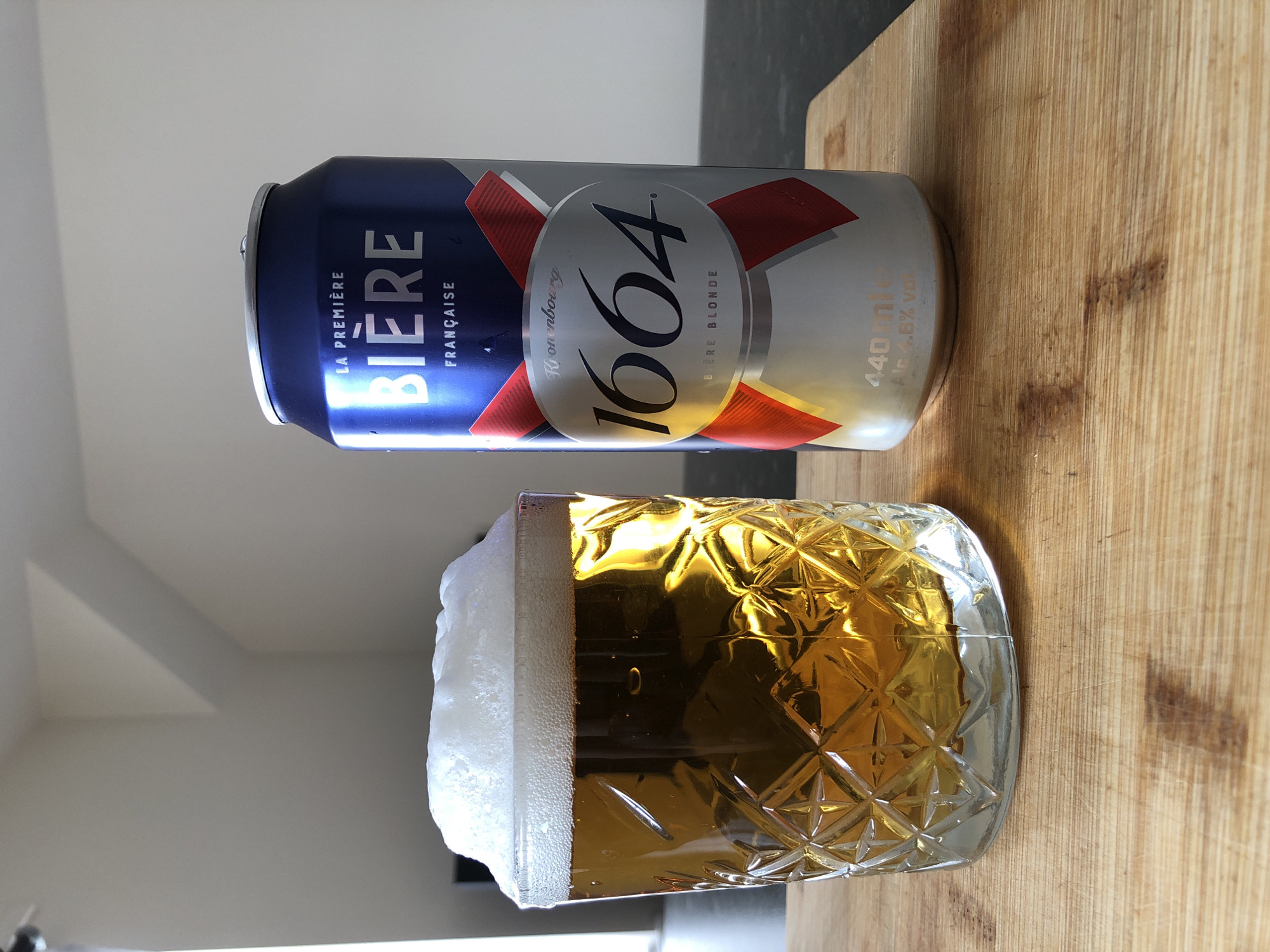 This media file depicts a beverage scene with a can and a glass of beer. The primary objects are a dark blue and silver aluminum can with red and white accents, and a clear glass filled with a golden-amber liquid topped with a thick white foam. The beer can is partially visible, showing text that includes "LA PREMIÈRE BIÈRE FRANÇAISE" and "1664 BIÈRE BLONDE". It also displays "440ml" and "Alc 4.6% VOL". The glass is intricately patterned, with a diamond-like texture, and the beer inside reflects light.
The setting appears to be indoors, with a plain white background visible behind the can and to the left of the glass. The objects are placed on a light-colored wooden surface, which takes up the right side of the image. The lighting suggests it is daytime, with natural light contributing to the reflections on the glass. No people are visible in the frame, and no specific event is taking place other than the presentation of the beer. The overall mood is neutral, focusing on the products themselves. The image is oriented vertically.