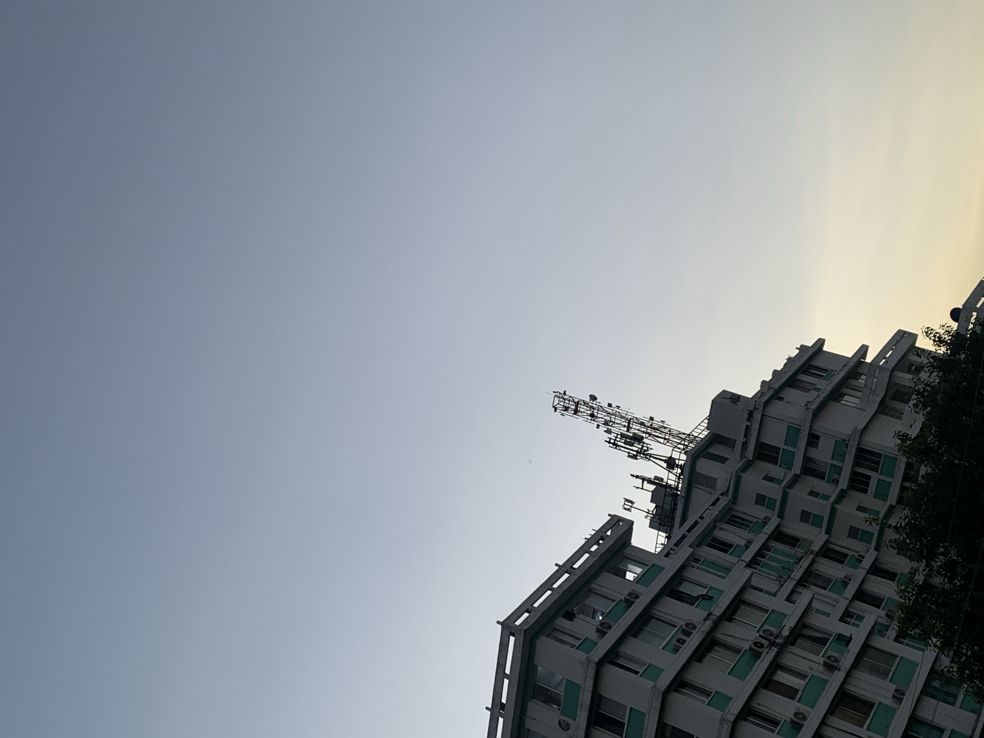 A low-angle shot reveals a tall, multi-story building on the right, equipped with numerous antennas and communication devices on its roof. The building features rows of windows and air conditioning units. Its angular design is prominent against the sky. To the left is a clear, light blue sky, transitioning to a faint yellow or orange hue near the top right corner, suggesting either dawn or dusk. A dark, leafy tree occupies the far-right edge. Based on the building's style and available evidence, the location is plausibly Dar es-Salaam, Tanzania. No people are visible.