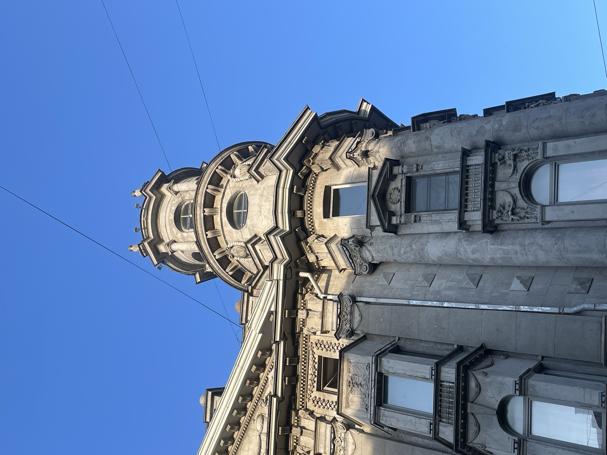 The image presents a striking, upward-looking view of a richly ornamented building facade against a clear, vibrant blue sky, indicative of daytime and good weather. The architecture suggests a grand, historic style, characteristic of cities like Saint Petersburg, Russia.
The primary subject is the building, crafted from what appears to be grey stone or concrete, showcasing elaborate architectural details. A prominent feature is a rounded turret or corner section, adorned with multiple circular windows and intricate relief carvings. The facade is layered with decorative elements, including detailed cornices, friezes, and moldings with sculptural accents, such as small figures resembling gargoyles. A distinctive checkerboard pattern can be observed along one of the horizontal bands.
The building features various window types, including rectangular windows with simple frames and at least one semi-circular or arched window. Fluted pilasters or columns are also visible, adding to the classical or Art Nouveau aesthetic. Sunlight illuminates the building from the left, creating shadows that emphasize the depth and texture of the architectural details. Several thin, dark wires crisscross the bright blue sky. There are no people or discernible text visible in the image.
