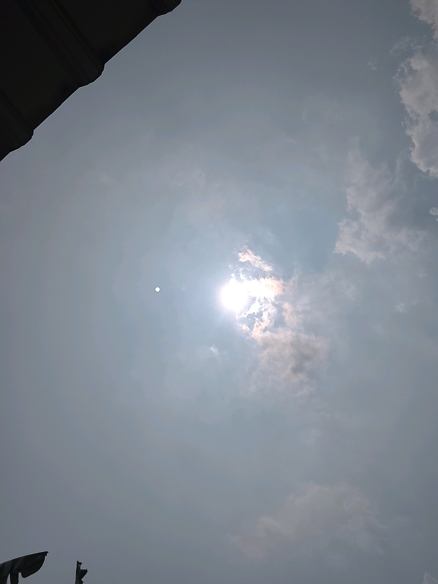 The image captures a view of the sky in Slawi, Indonesia, dominated by clouds and the sun. A portion of a dark, corrugated roof is visible in the upper left corner, suggesting an elevated vantage point. The sun is a bright, diffused orb peeking through scattered clouds, creating a hazy glow. Several clouds are present, some appearing white and wispy, while others are darker and more dense, suggesting varying atmospheric conditions. In the lower part of the frame, two dark, abstract shapes are visible, possibly flags or banners, silhouetted against the sky. The overall impression is one of a cloudy day with the sun attempting to break through.