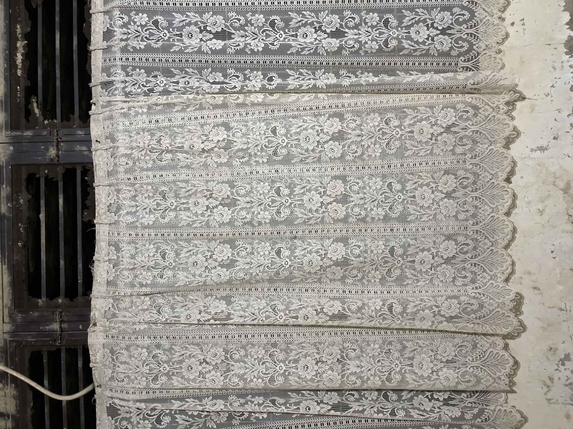 The image displays a close-up of a white lace curtain, draped and folded. The curtain features a repeating floral pattern with delicate, intricate designs. It is hung over what appears to be a dark, barred window or vent. The background wall on the right is textured and painted white, with visible imperfections and shadows, suggesting an interior setting. The lighting appears to be artificial, casting shadows that highlight the folds of the lace. The overall impression is one of a somewhat neglected or rustic environment, with the delicate lace providing a stark contrast to the rougher textures of the surroundings. There is no text visible in the image. The location context provided is Kota Kuala Simpang, Indonesia. The image captures a still, quiet scene with no people or discernible activity.