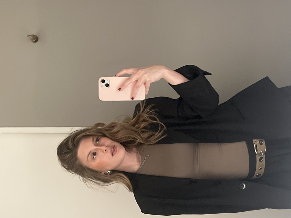 A woman takes a selfie. She is wearing a black blazer, a tan shirt, and a beige belt. Her hair is wavy and shoulder-length. She holds a pink phone in her left hand. The background is a gray wall with a metal knob. The woman is looking at the camera with a slight smile.