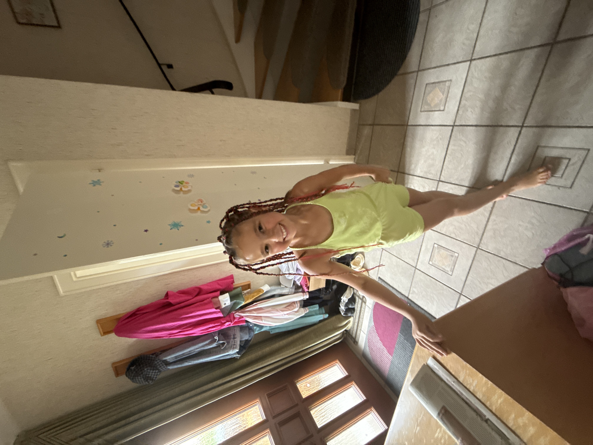 A young girl with braided hair is standing on a tiled floor in an indoor entryway. She is wearing a light green romper and smiling at the camera. To her left is a wooden door with glass panels, and to her right is a staircase. Coats and clothing are hanging on hooks on the wall behind her. A table with a newspaper on it is in the foreground. The scene appears to be a residential entryway, possibly in Bonn, Germany, given the context provided.