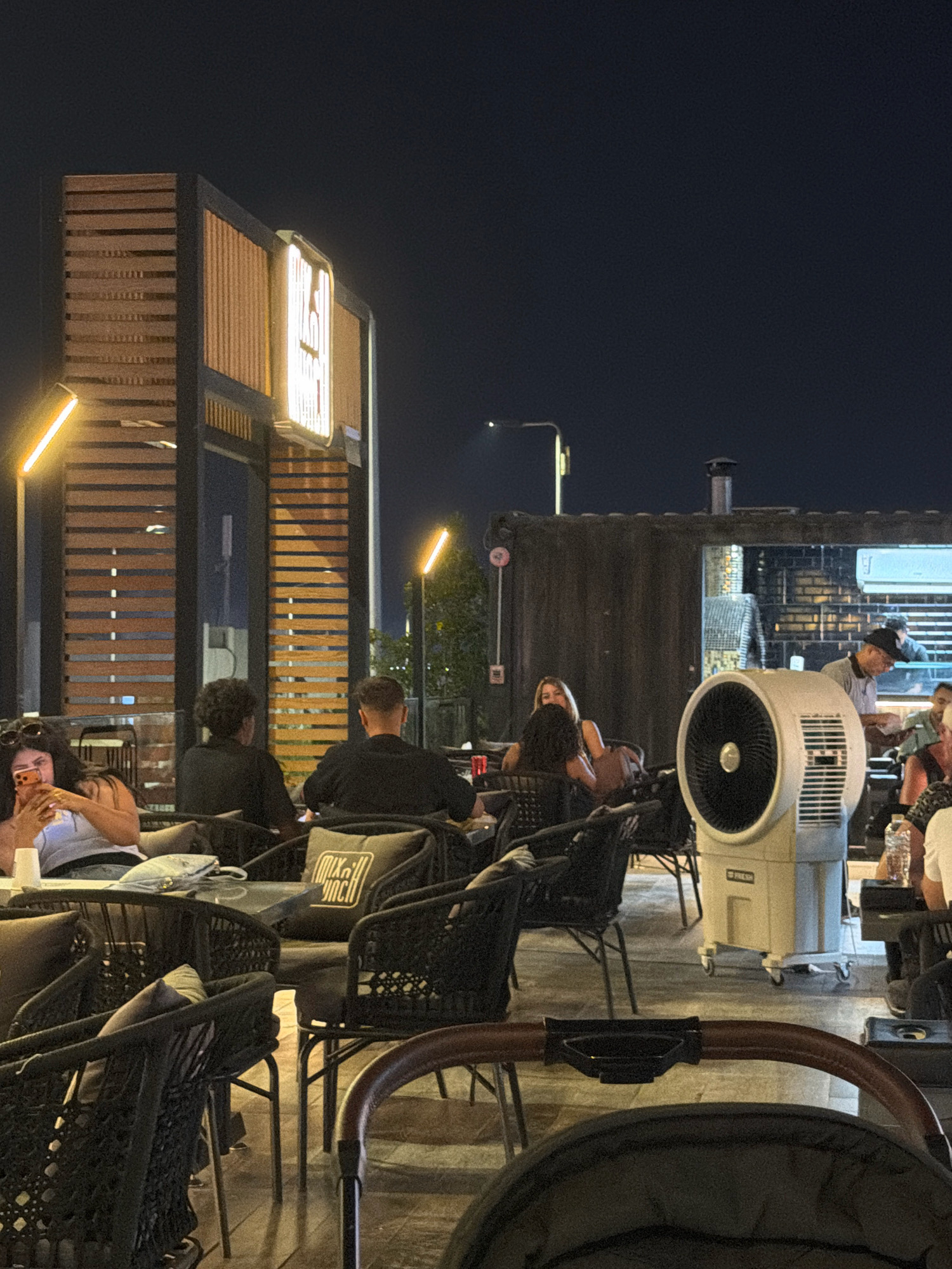 The image depicts an outdoor seating area at night, likely a restaurant or cafe, in Sharm El Sheikh, Egypt. Several individuals are seated at tables, some engaged in conversation, others looking at their phones. A prominent white industrial-style fan stands to the right of the frame, suggesting a warm climate. The architecture features wooden slats and modern lighting. In the background, a building with exposed brick and a bright light fixture is visible. The overall atmosphere is casual and social.