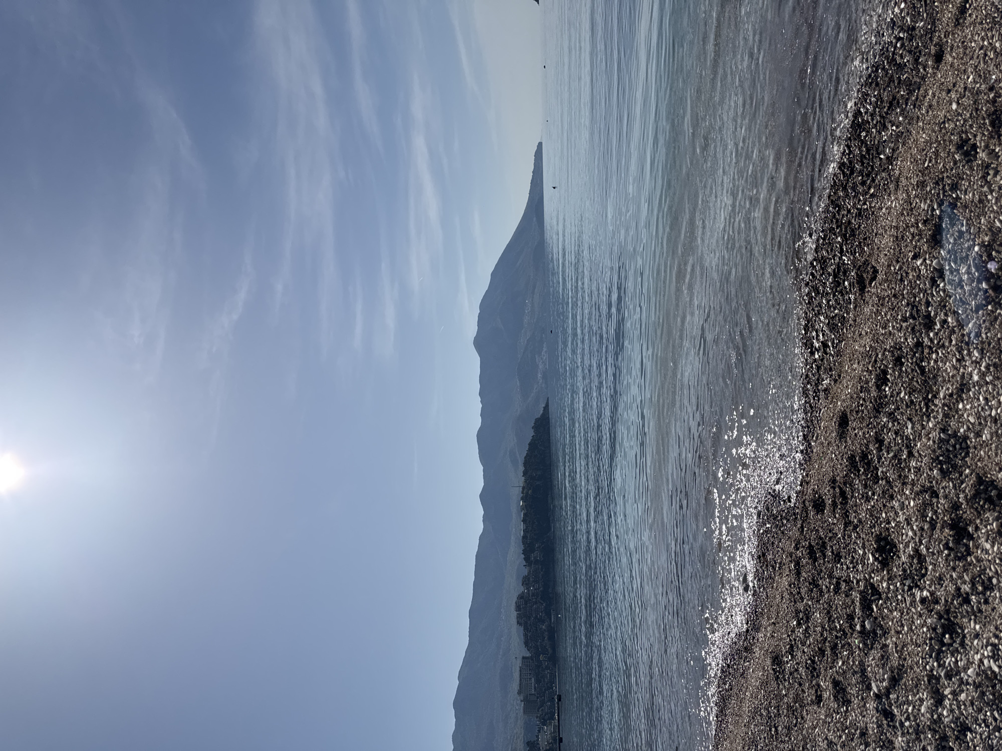 The image shows a beach scene in Budva, Montenegro. The Adriatic Sea meets a dark pebble beach in the foreground, with sunlight reflecting off the water. In the background, hazy mountains line the coast. The sky is pale blue with wispy clouds, and the sun shines brightly on the left side of the frame. Buildings are visible at the base of the mountains.