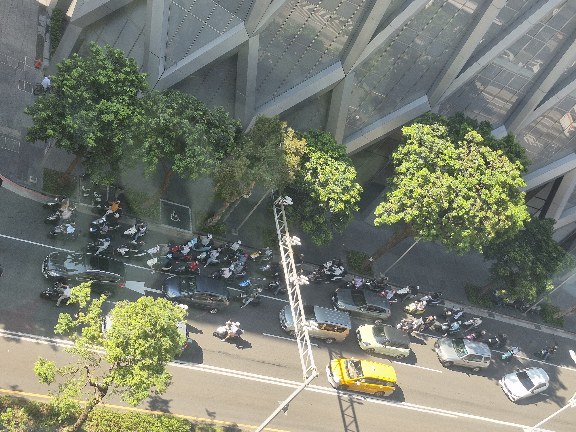 The image is an aerial view of a street scene in Taipei, Taiwan, likely taken from a high-rise building. The scene is dominated by a multi-lane road filled with various vehicles, including a yellow taxi, several cars, and a multitude of scooters and motorcycles. People are seen riding or dismounting these vehicles. A modern glass-fronted building with geometric architectural features stands to the left of the road, casting long shadows. Lush green trees are interspersed along the roadside and in front of the building, some of which are casting dappled shadows on the road. The overall impression is a busy urban environment with typical street life in Taipei.