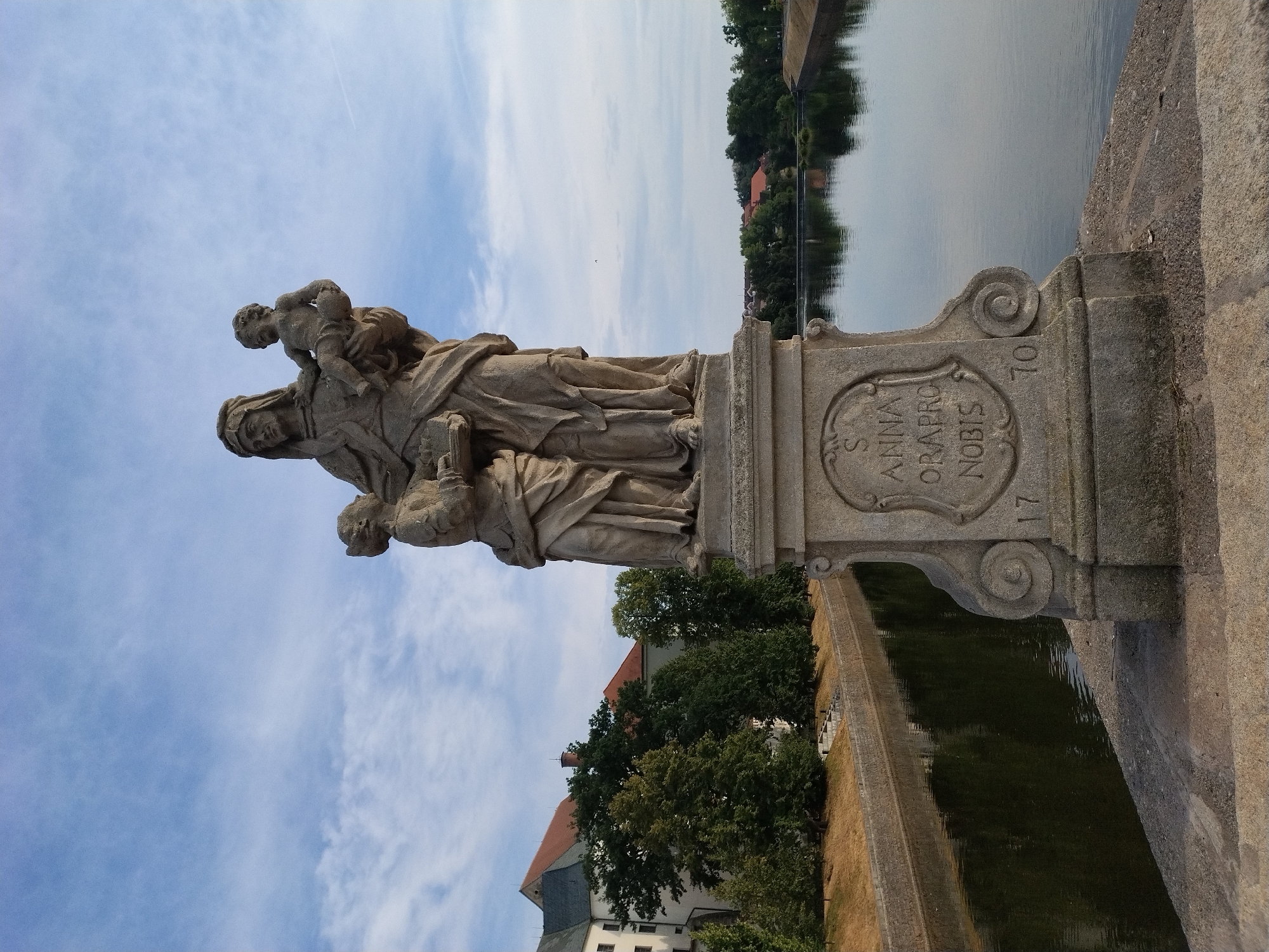 The image depicts a stone sculpture of Saint Anne holding the infant Jesus and another child. The sculpture is mounted on a pedestal with inscribed Latin text "S ANNA ORA PRO NOBIS" and the date "1770". It is situated on a stone bridge spanning a body of water, likely a river, with a canalized embankment visible. In the background, there is a building with red tiled roofs and trees, suggesting an urban or semi-urban environment. The sky is partly cloudy. The sculpture is located on the Písek Stone Bridge (Kamenný most v Písku) in Písek, Czechia.