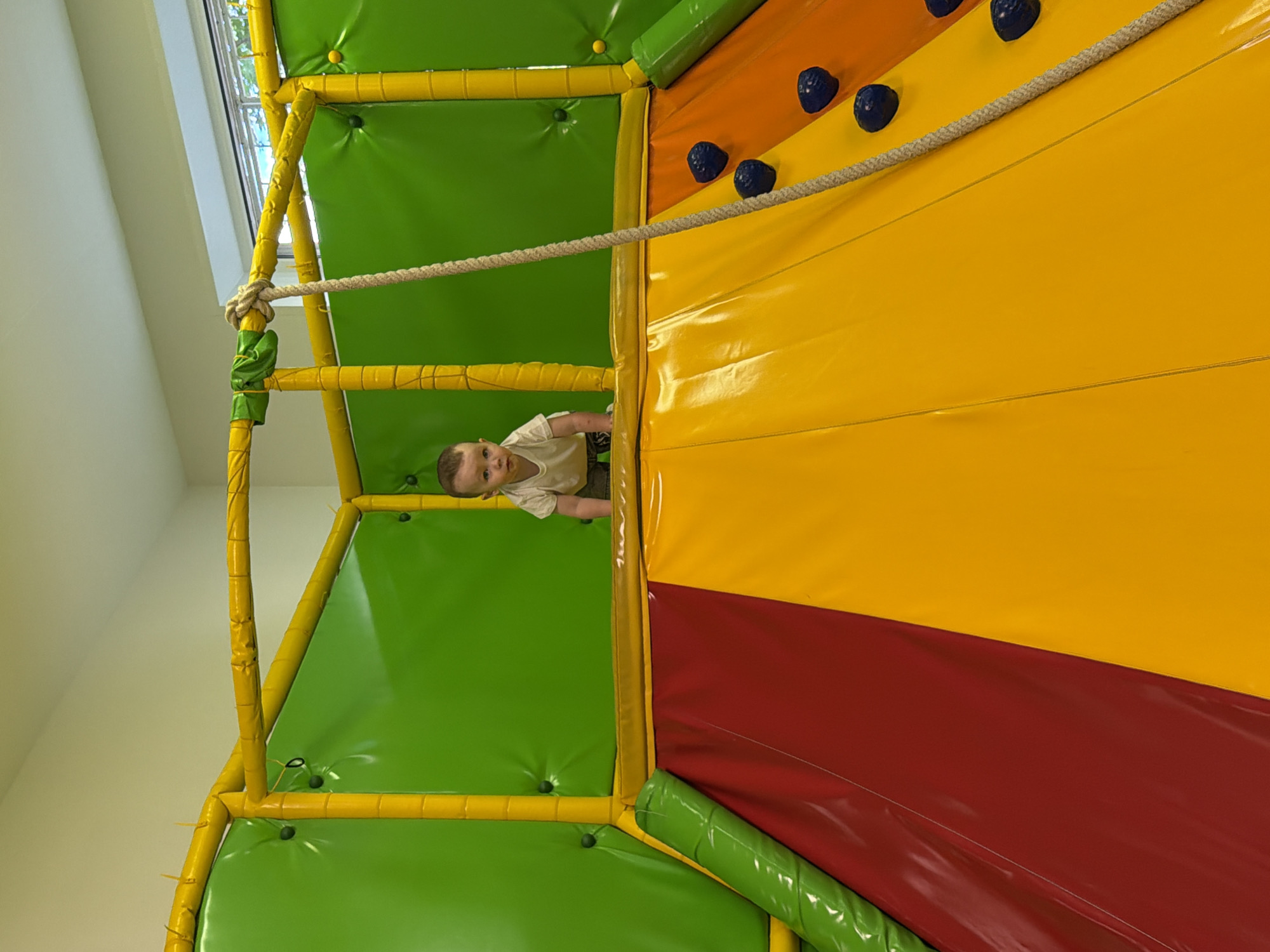 A baby in a white t-shirt and shorts is crawling on a soft play structure in a room. The structure is made of green and yellow padded material, with a red and orange slide in the foreground. A rope hangs from the top of the structure. The scene appears to be indoors, possibly in a children's play center. The image does not contain any identifiable landmarks or information that would allow for its location in Chaikovsky, Russia.