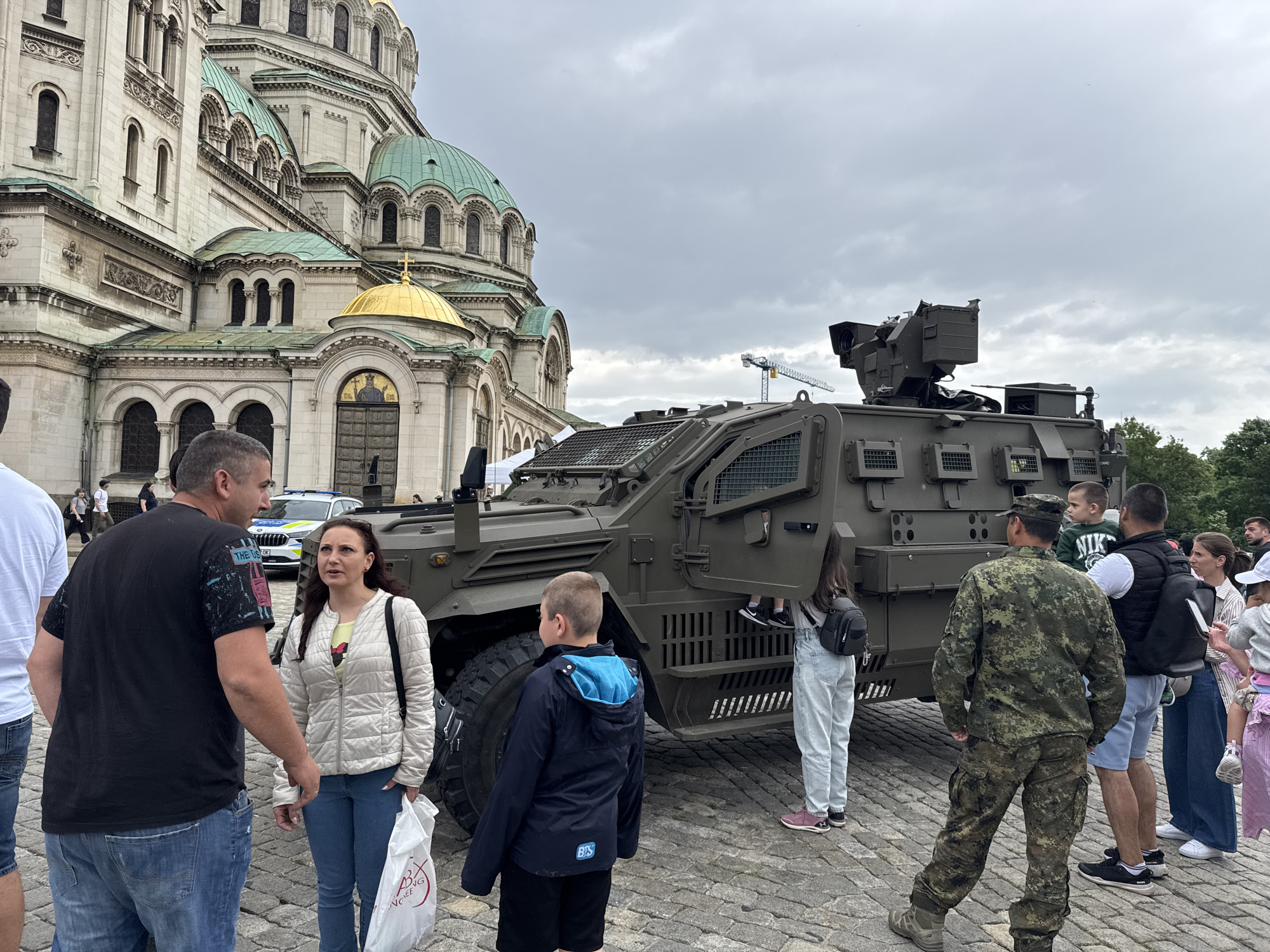 In an outdoor scene on a cloudy day, a group of people are gathered around a military vehicle in the city of Unknown, Unknown. The vehicle is olive green and appears to be an armored personnel carrier. It is parked on a cobblestone surface. A large cathedral-like building with multiple green and gold domes stands in the background, partially obscured by the vehicle and the people. The individuals present include adults and children, some of whom are standing near the vehicle, while others are conversing with each other. One man is wearing military camouflage. A white police car is visible in the background behind the group. The scene suggests a public display or exhibition of military equipment.