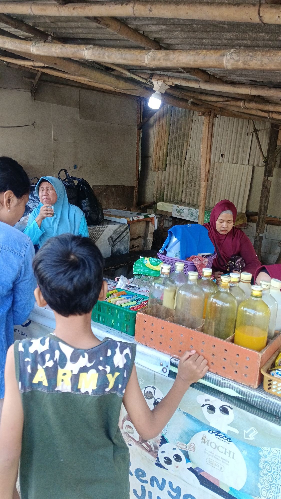 In Tajurhalang, Indonesia, a street vendor with a maroon hijab serves drinks from glass bottles to customers in an open-air stall. The stall is constructed from bamboo and corrugated metal sheeting. A young boy in a green sleeveless shirt with "ARMY" written on the back faces the vendor, his left hand resting on the counter. To the left, a woman in a blue hijab drinks from a glass. The counter displays bottles of orange liquid, a green basket of colorful items, and a package labeled "Alice MOCHI." The scene suggests a local market or roadside food stall.