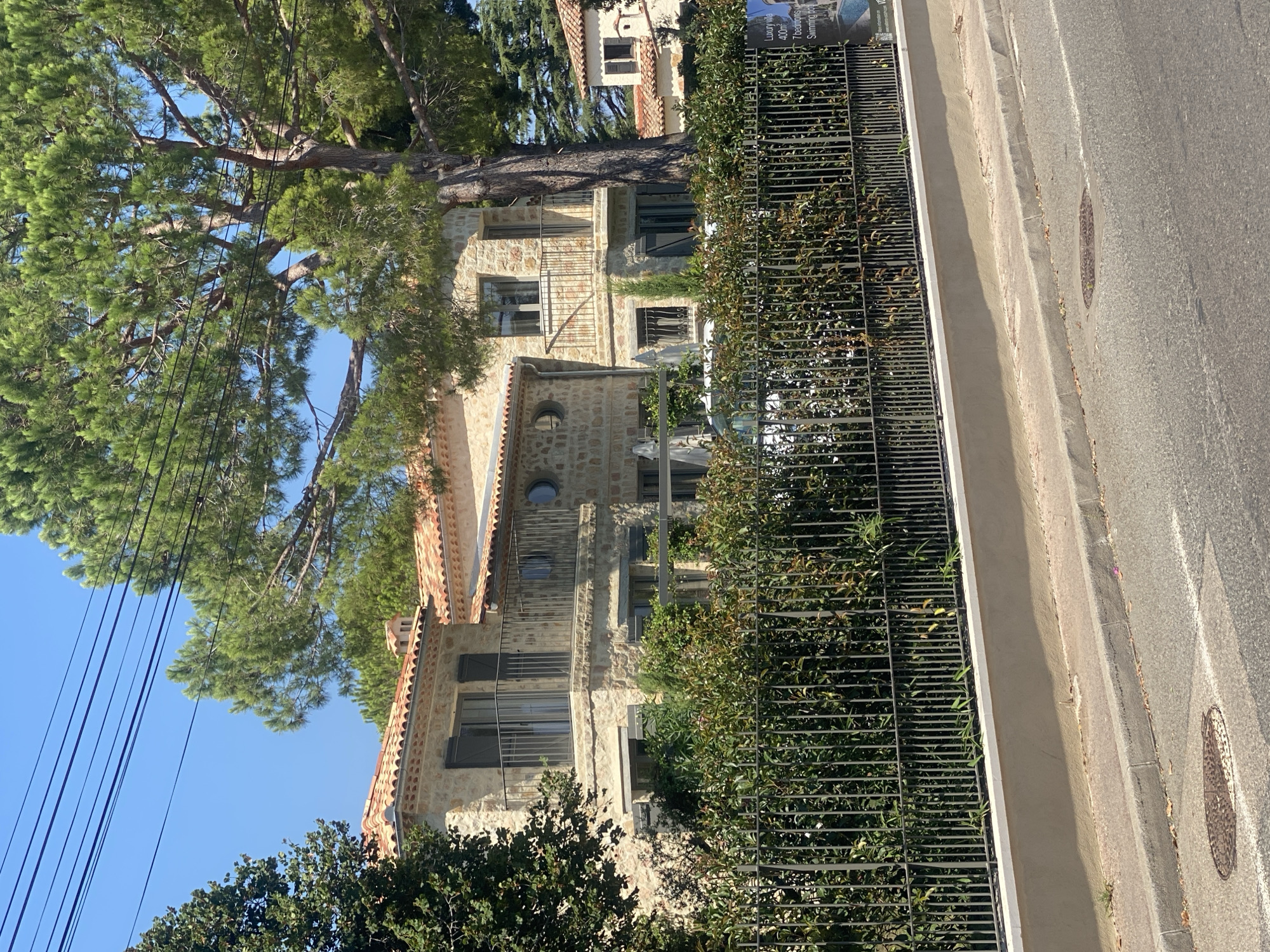 This image depicts a large, multi-story stone villa situated in Antibes, France. The villa features a terracotta tiled roof, multiple balconies with black metal railings, and dark shutters on the windows. It is surrounded by lush green foliage, including a prominent large pine tree to the right of the house. A black metal fence separates the property from a paved street in the foreground. A white wall runs along the base of the fence. The sky is clear and blue. To the far right, a portion of another building, also with a terracotta roof, is visible. There are power lines visible against the sky. A sign is partially visible behind the fence on the right side of the image, with text that appears to indicate real estate information, including "Luxury villa 400m², 7 bedrooms, Swimming pool." No people are visible in the scene. The overall impression is one of a well-maintained, upscale residential property in a sunny climate.