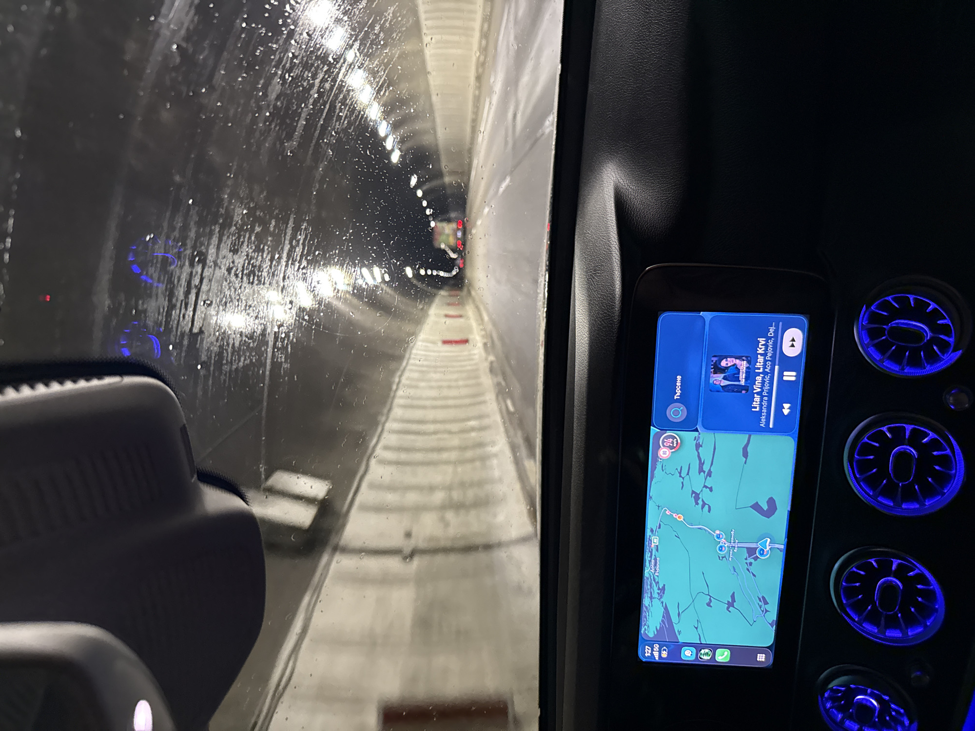 The image shows a driver's perspective inside a vehicle traveling through a tunnel. The tunnel walls are white and illuminated by rows of lights. The windshield is wet with water droplets. A dashboard screen to the right displays a map and music information. The map shows the location of Dolna Vasilitsa, Bulgaria. The music playing is "Litar Vina, Litar Krvi" by Aleksandra Prijovic, Aco Pejovic, and Dej. To the right of the screen, there are circular vents with blue illumination.