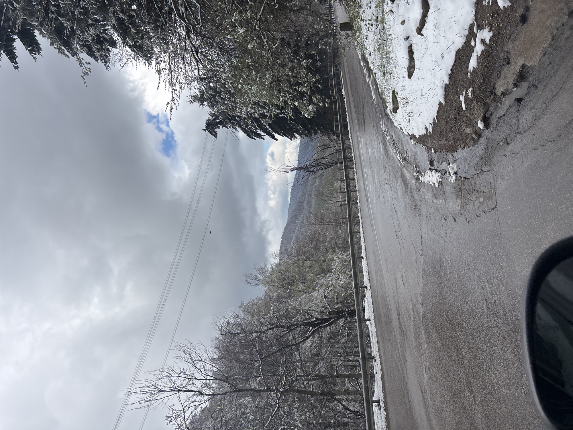 The image shows a wet asphalt road curving slightly to the right. On the right side of the road, there is a shoulder with patches of snow. A metal guardrail runs along the left side of the road. Trees covered with snow line both sides of the road. In the background, a mountain is visible, partially obscured by clouds. Overhead, power lines stretch across the sky. The sky is overcast with dark, heavy clouds. A small patch of blue sky is visible through a break in the clouds. The image seems to have been taken from inside a car. It's likely from the area near the city of Barziya in Bulgaria, given the presence of snowy conditions and mountainous terrain.
