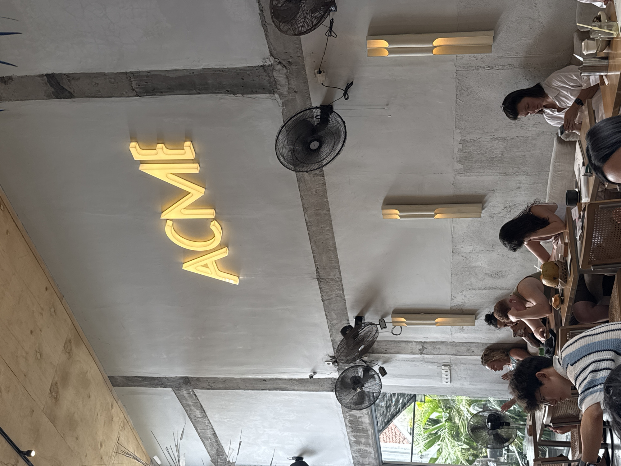 The image shows the interior of a restaurant, reportedly in North Kuta, Indonesia. The ceiling is visible, featuring concrete beams, fluorescent lighting fixtures, and several wall-mounted electric fans. A large illuminated "ACME" sign is prominently displayed. Below the ceiling, customers are seated at tables and appear to be engaged in various activities, likely dining and socializing. The image provides a glimpse of the restaurant's ambiance and the customers present. The scene includes a window with greenery visible outside.