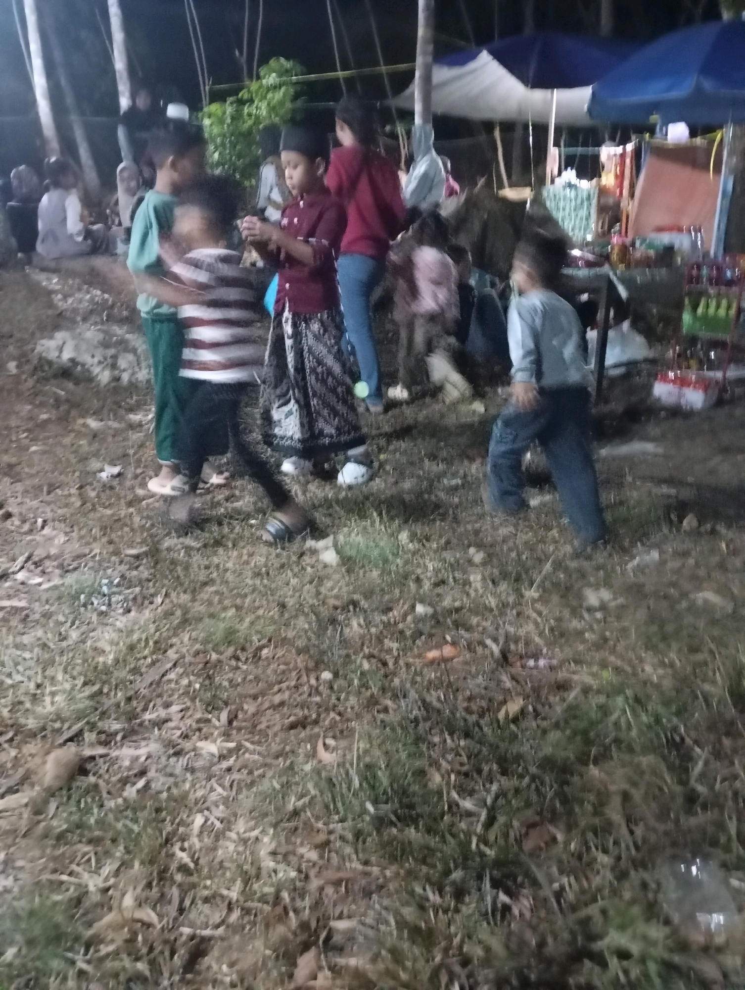 A nighttime outdoor scene, possibly a market or gathering, is captured in Ketepung, Indonesia. In the foreground, the ground is covered with dry grass, leaves, and scattered debris. Several children are present. One child, wearing a striped shirt and dark pants, is in motion. Another child, in a maroon shirt and patterned pants, stands facing left, holding something in their hands. To the left, a child in a green shirt is partially visible. Further back, other individuals are present, some seated. In the background, there are tents with blue and white canopies, suggesting market stalls or temporary structures. Objects, possibly merchandise, are visible on shelves behind some of the tents. The lighting is dim, with artificial lights illuminating the scene.
