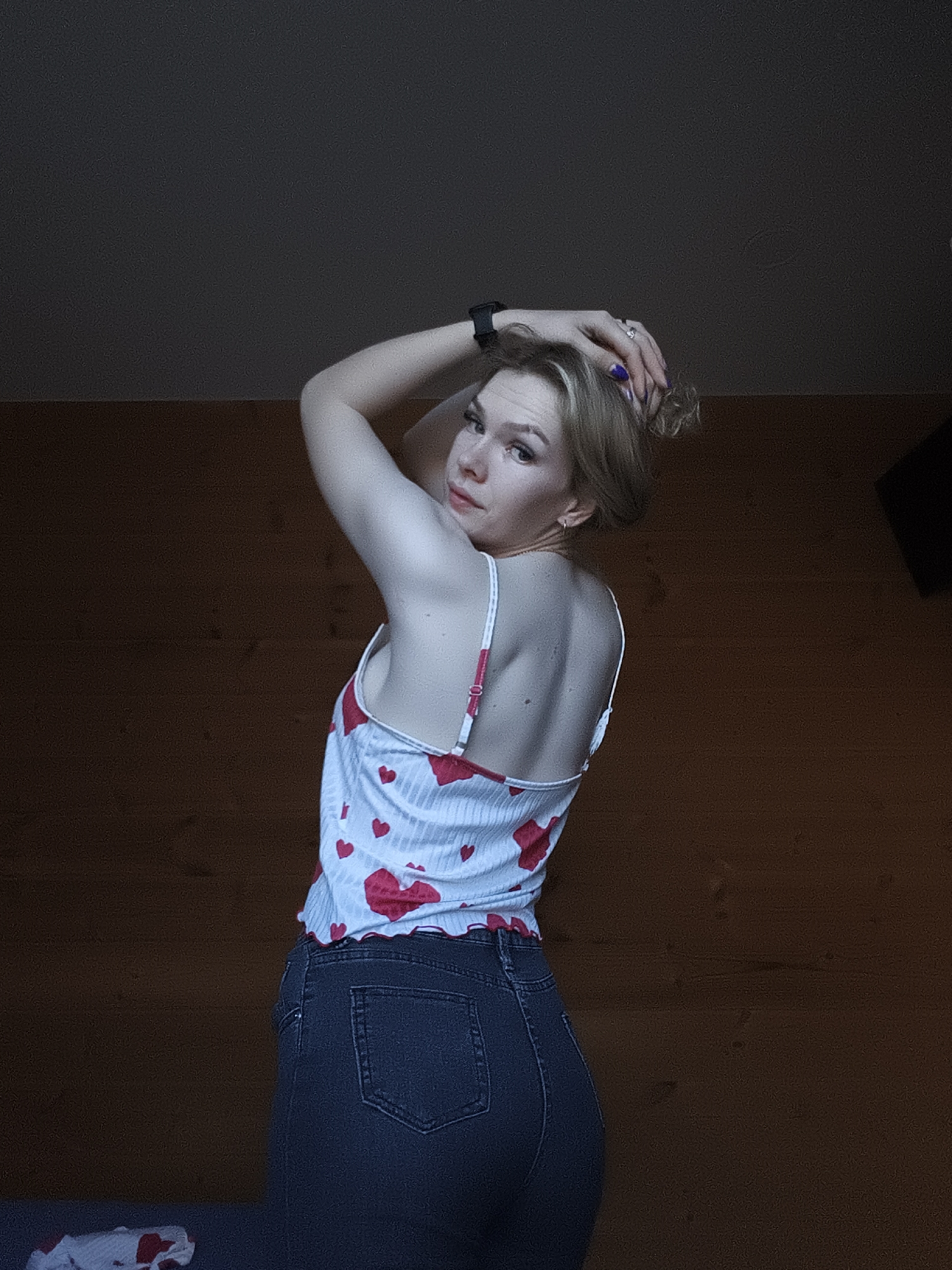 A fair-skinned woman is posing indoors, turned towards the viewer. She is wearing dark blue jeans and a white, heart-patterned camisole. Her right arm is raised, with her hand touching her hair, and she is wearing a dark-colored watch. She is looking at the viewer. The background consists of a dark wooden wall and ceiling, with a dark object hanging in the top right. The location is unknown but is not indicated as Русский Вожой, Russia.