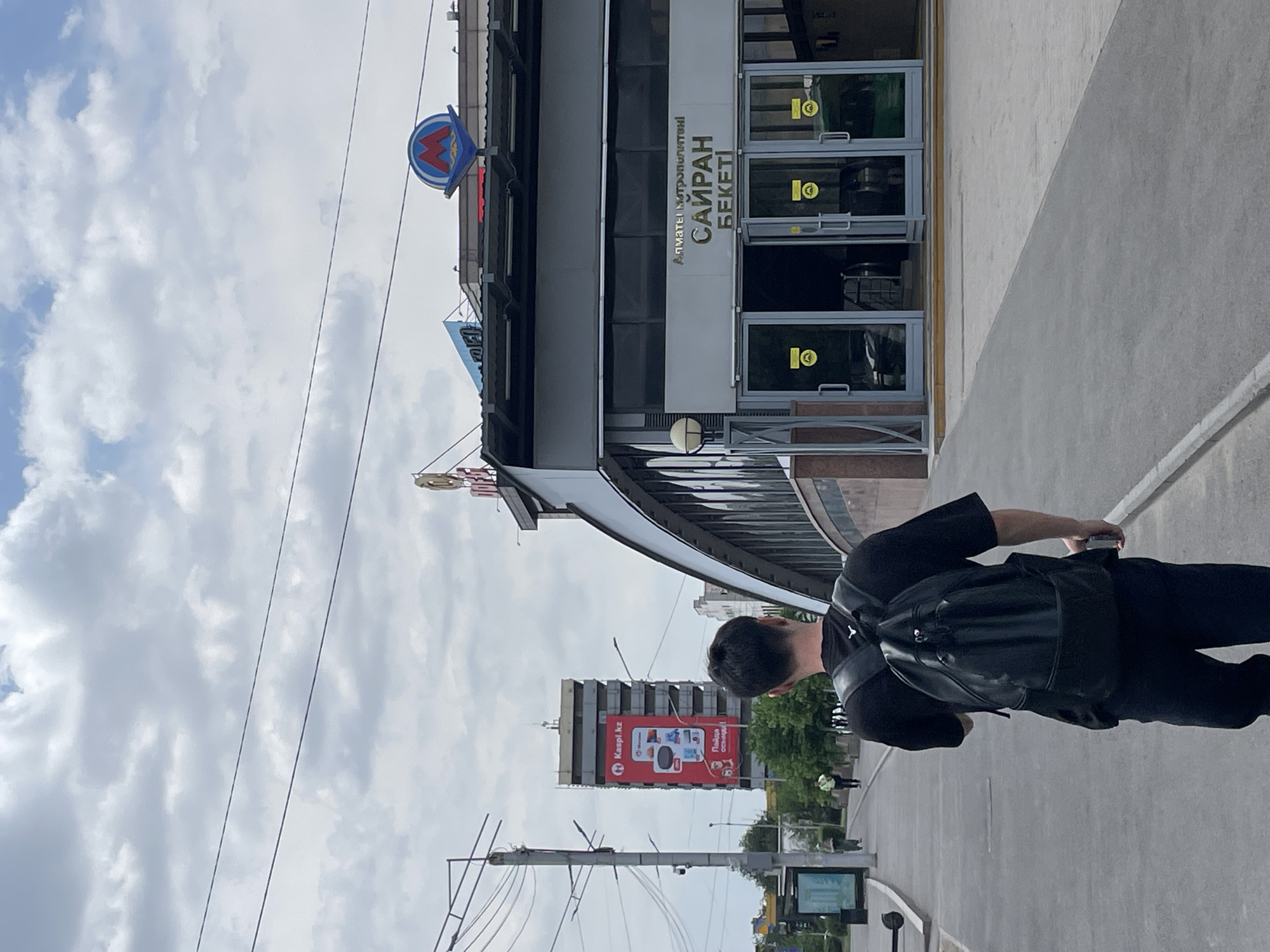 The image shows a person wearing a black t-shirt and black pants, with a black backpack. They are standing on a sidewalk next to a building. The building has a metro station sign with the letter "M" in a red circle. Above the entrance, there is text in Cyrillic that reads "АЛМАТЫ МЕТРОПОЛИТЕНІ" and "САЙРАН БЕКЕТІ". This translates to "Almaty Metro" and "Sairan Station". The building appears to be modern with glass doors. In the background, there is a tall building with a large red billboard that has text in Cyrillic and Latin letters, including "Kaspi.kz". The sky is cloudy and appears to be daytime. The scene is likely in Almaty, Kazakhstan, given the text on the metro sign and billboard.