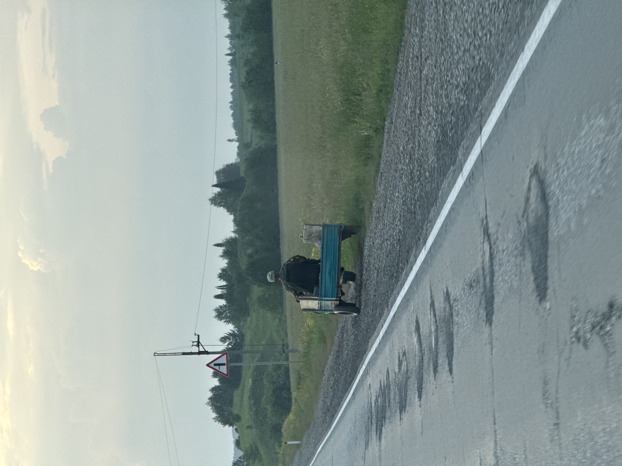 A man wearing a camouflage jacket and cap is sitting on a small, blue, two-wheeled trailer. The trailer is attached to the back of a slow-moving vehicle, not fully visible, on a gravel shoulder next to a paved road. The road has some potholes. The man is facing away from the camera. The environment is rural, with a field of green grass to the right of the road and trees on a gentle slope in the distance. A triangular road sign indicating a fork in the road is visible to the left of the road. The sky is overcast and pale. The scene appears to be captured from a moving vehicle.