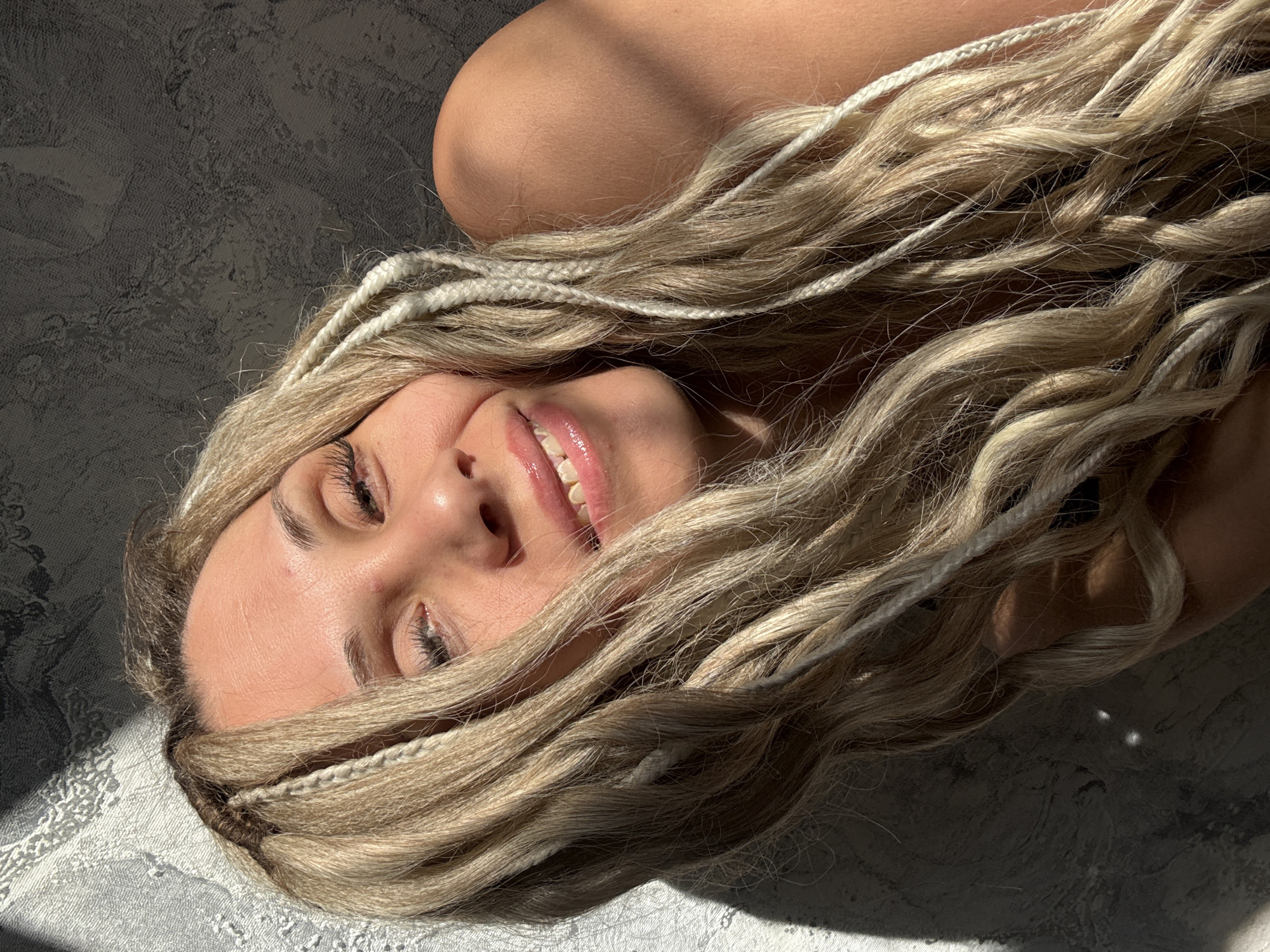 A woman with blonde and white braided hair lies on a grey surface, smiling slightly with her eyes partially closed. Light is cast across her face and hair. The location is unknown, and there is no evidence to suggest it is in Rusanovo, Russia.