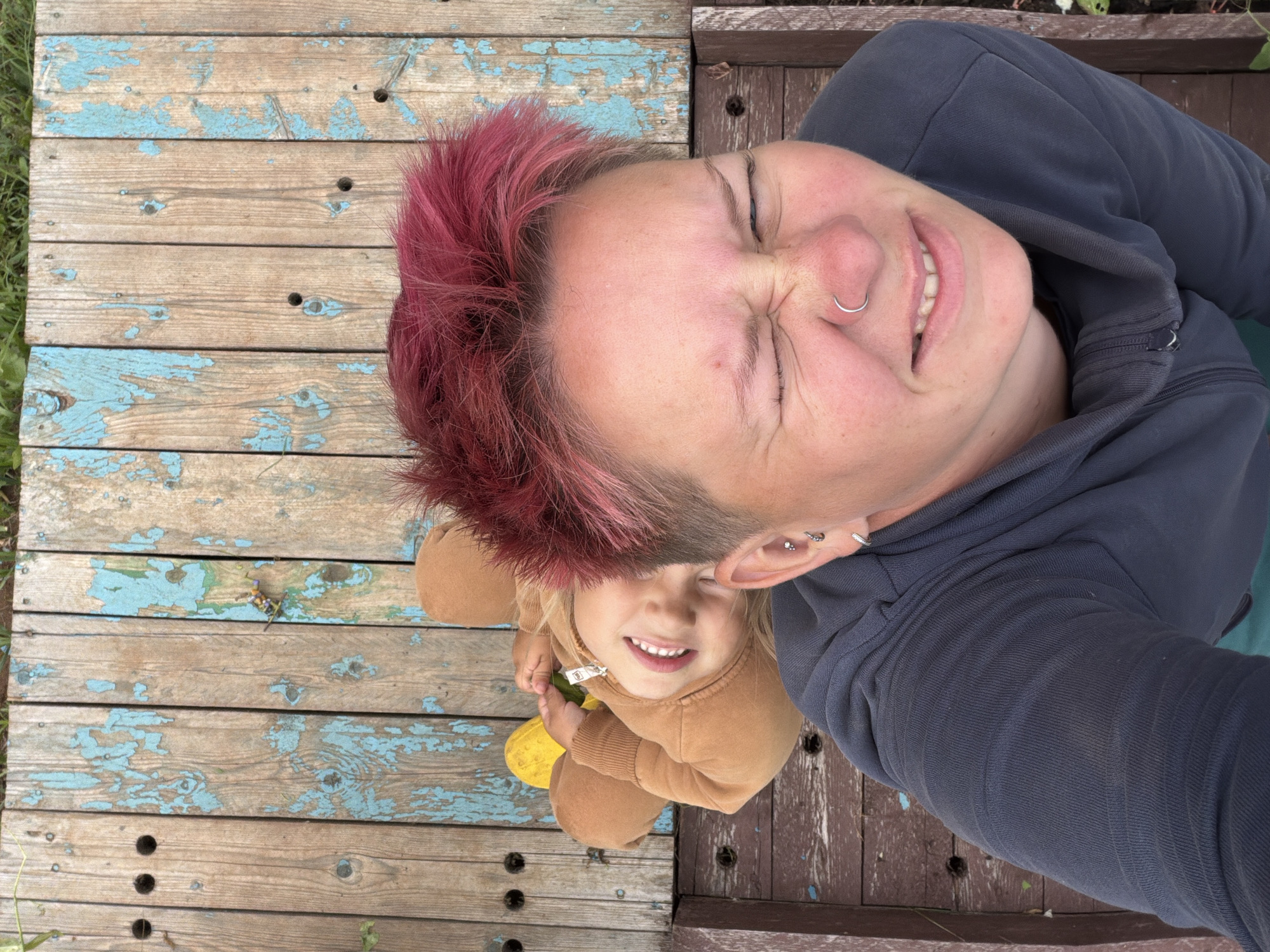A selfie taken from a high angle shows two people lying on a weathered wooden surface. The person in the foreground, with short, bright pink hair and a nose ring, is closing their eyes tightly and scrunching their face. They are wearing a dark grey hooded sweatshirt. Behind them, a young child in a tan hooded jumpsuit smiles widely, with their head tilted back. The wooden surface appears to be made of planks with peeling blue paint and visible grain. The background consists of more weathered wood, with hints of green foliage at the top and bottom edges. The scene suggests an outdoor setting, possibly a park or backyard. The location is identified as Chaikovsky, Russia.