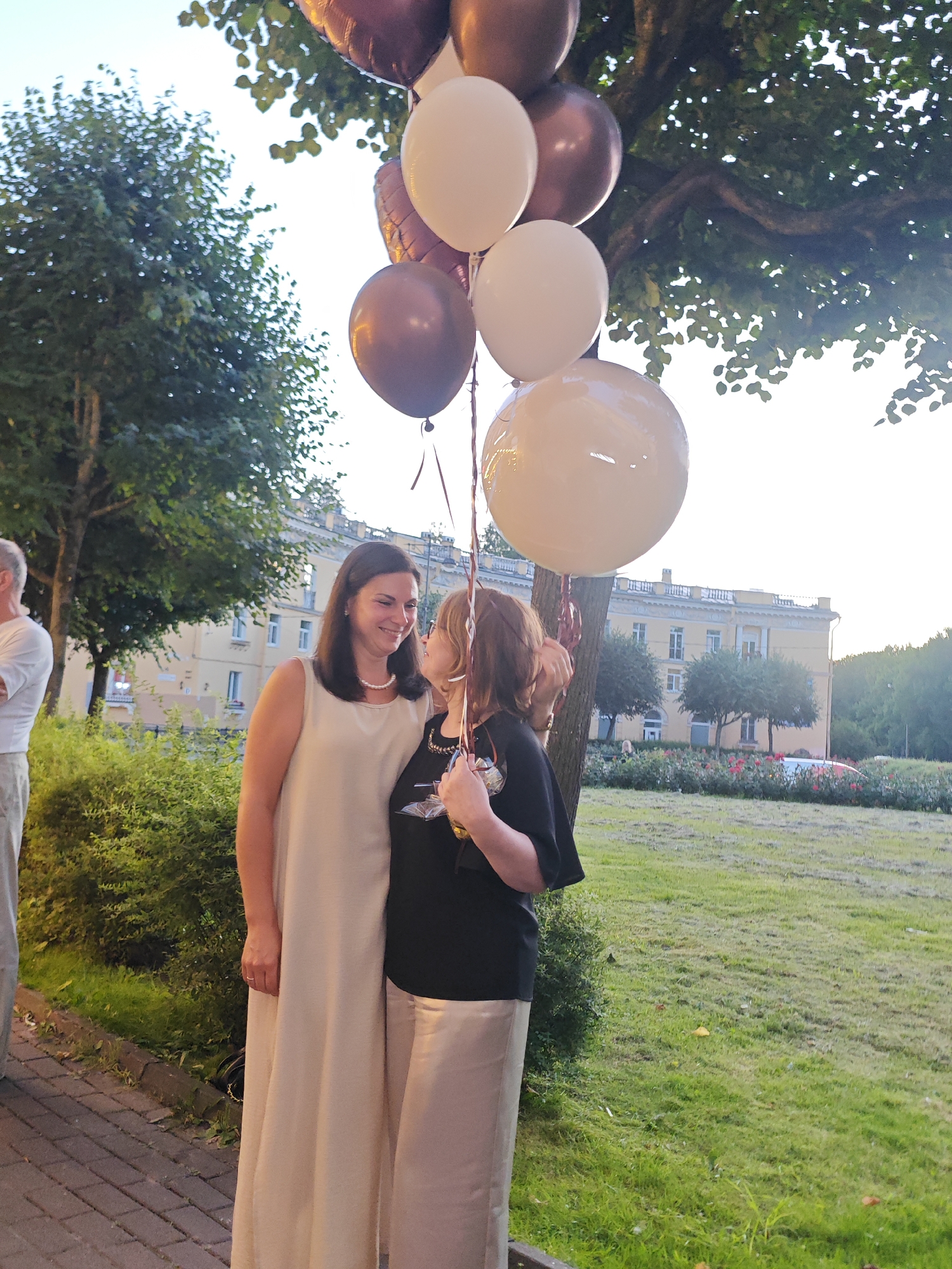 Two women are standing in front of a building in Pushkin, Russia. The woman on the left is wearing a long, cream-colored dress and a pearl necklace. She is smiling and looking at the woman on the right. The woman on the right is wearing a black t-shirt and light-colored pants. She is holding a drink and looking at the woman on the left. Both women are surrounded by balloons of various colors, including brown and cream. In the background, there is a building with a light-yellow facade and a red-tiled roof. The building is surrounded by trees and a green lawn. The sky is a pale blue with some clouds. The overall scene suggests a festive outdoor gathering.