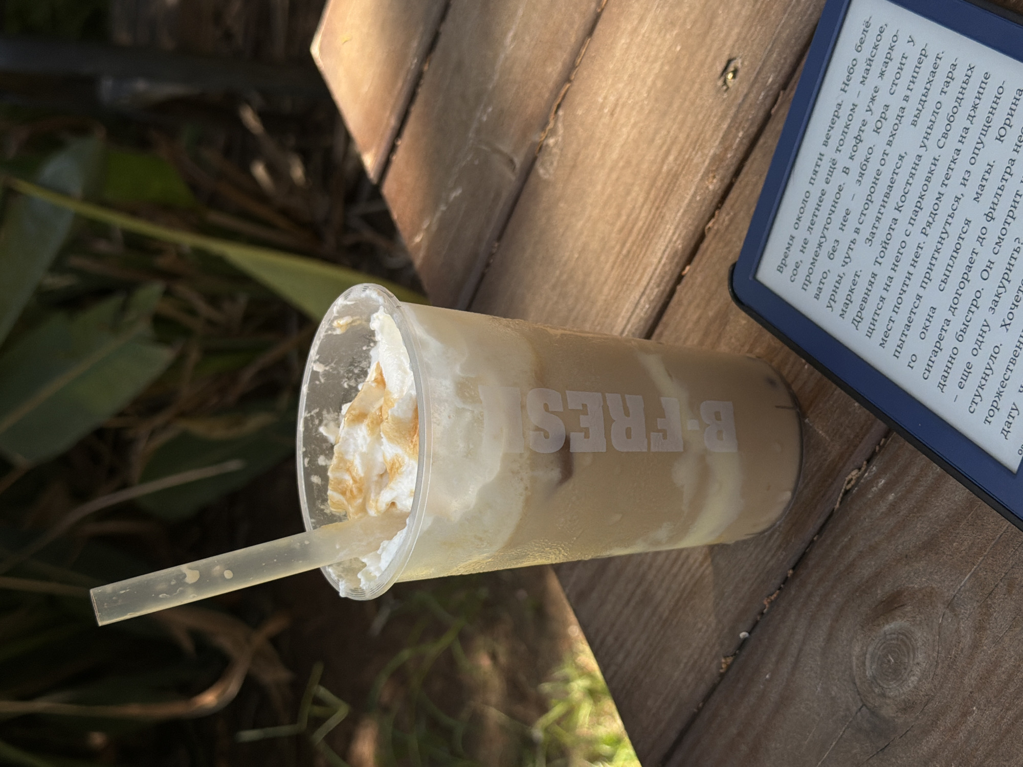 A clear plastic cup filled with a layered iced beverage, topped with whipped cream and a caramel drizzle, sits on a wooden table outdoors. A translucent straw extends from the cup. In the foreground, a blue e-reader displays text. The background consists of blurred greenery, suggesting an outdoor cafe or patio setting. The image is taken from a slightly elevated angle, looking down at the table. The lighting indicates it is daytime, with sunlight casting shadows. The text on the cup appears to read "B-FRESH". The overall scene suggests a moment of relaxation and reading.
